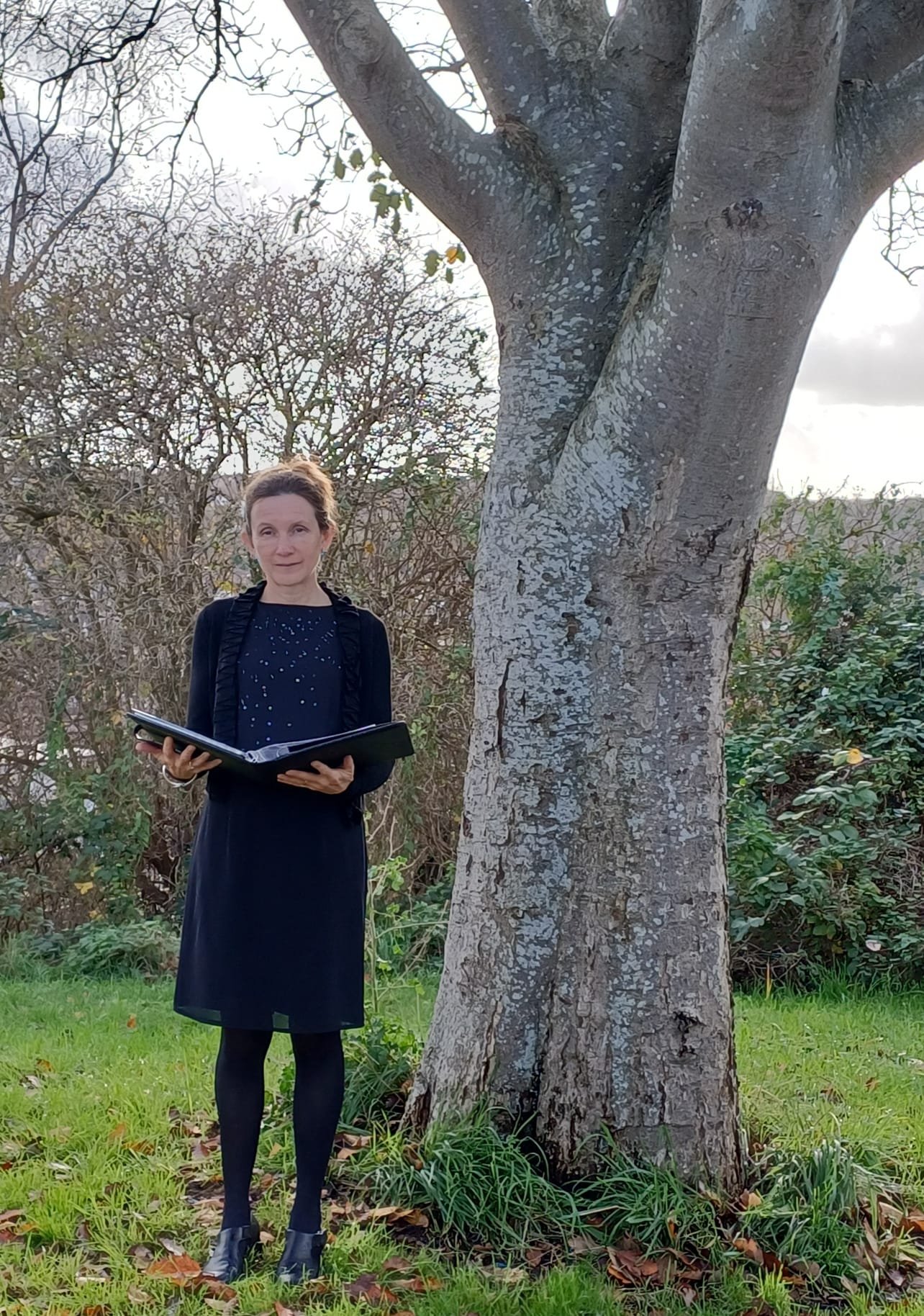 Rachel Peatfield, Celebrant - standing by tree with black portfolio folder at outdoor memorial