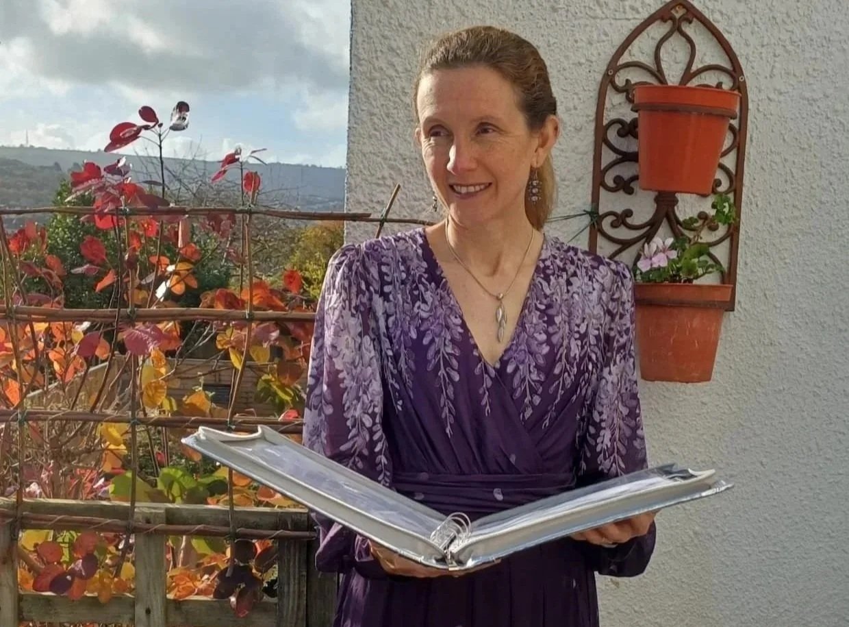 Rachel in purple dress with silver ceremony folder, golden leaves and sky in background