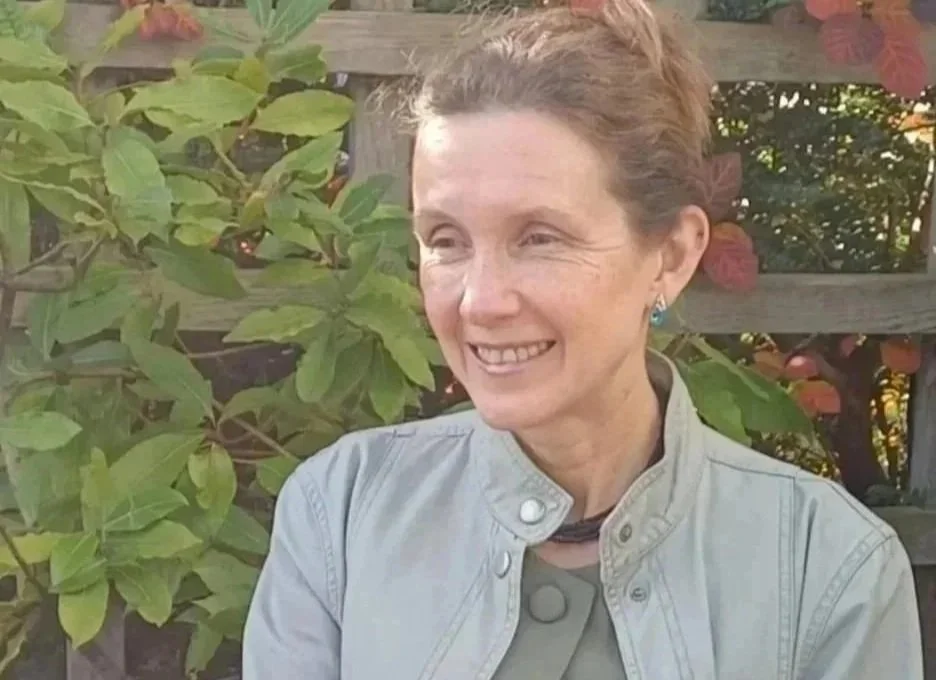 Rachel in grey jacket and green-grey trouser suit with green leafy background