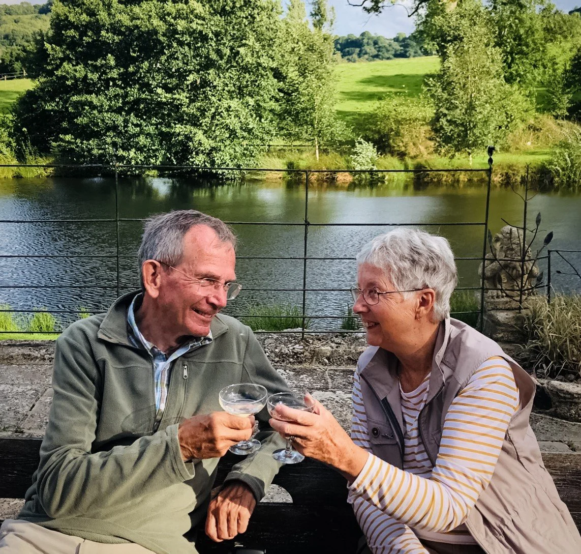 Older man and woman clinking champagne glasses by a lake with fields and trees in background.
