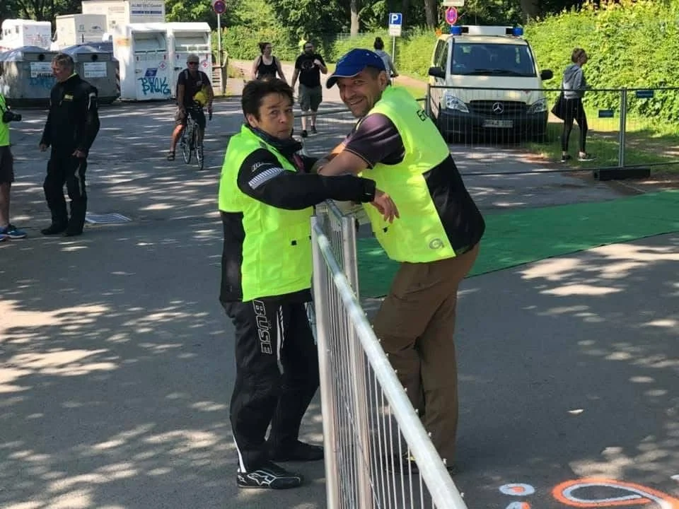 Zwei Menschen in neon gelben Sicherheitswesten unterhalten sich an einem metalldrahtgitterzaun in einer öffentlichen Parkanlage. Im Hintergrund sind weitere Personen, ein Autos, ein Fahrrad und Grünflächen sichtbar.