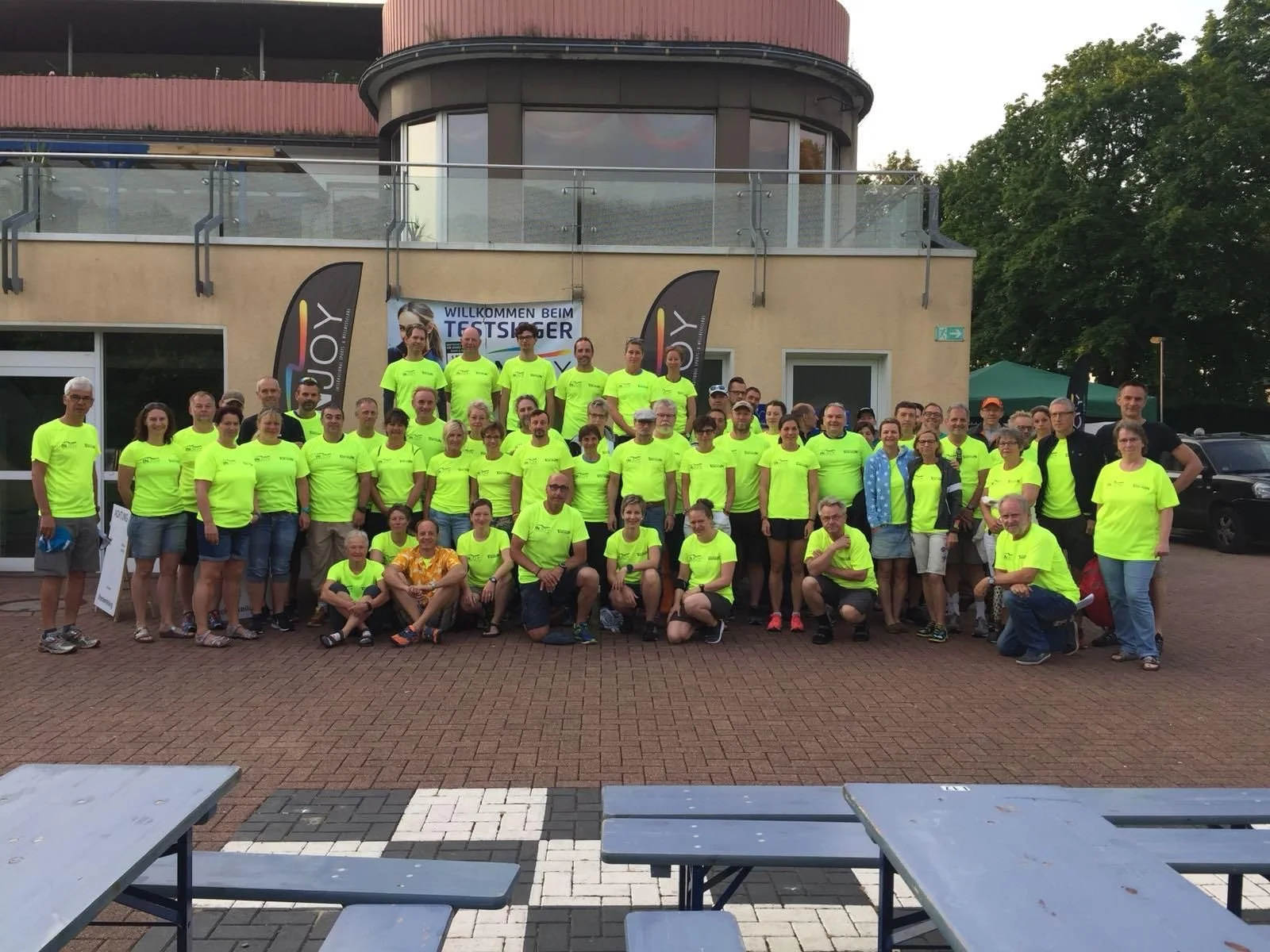 Eine große Gruppe von Menschen trägt neon gelbe T-Shirts und posiert für ein Gruppenfoto vor einem Gebäude mit einem Banner im Hintergrund, das Willkommen beim Testsieger sagt.