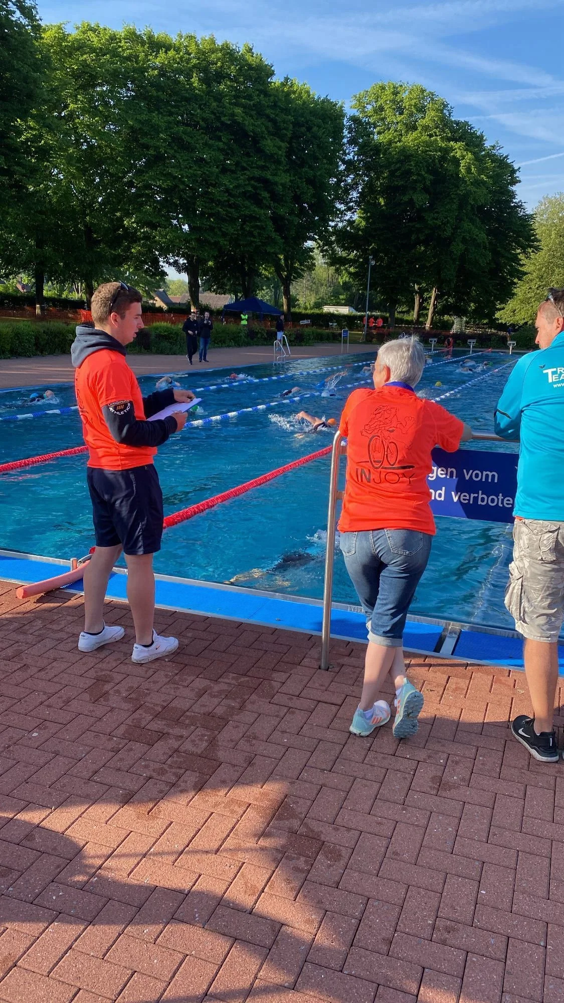Menschen beobachten ein Schwimmtraining im Freien mit Aquathermline. Zwei Frauen und ein Mann stehen am Beckenrand. Mehrere Schwimmer schwimmen im Wasser, umgeben von Bäumen und einem blauen Himmel.