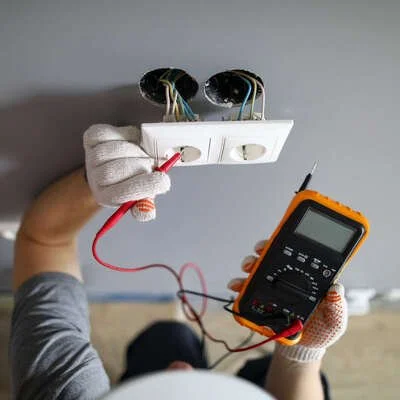 Une personne effectue un dépannage électrique en utilisant un multimètre pour tester des prises électriques, tout en portant des gants de protection.