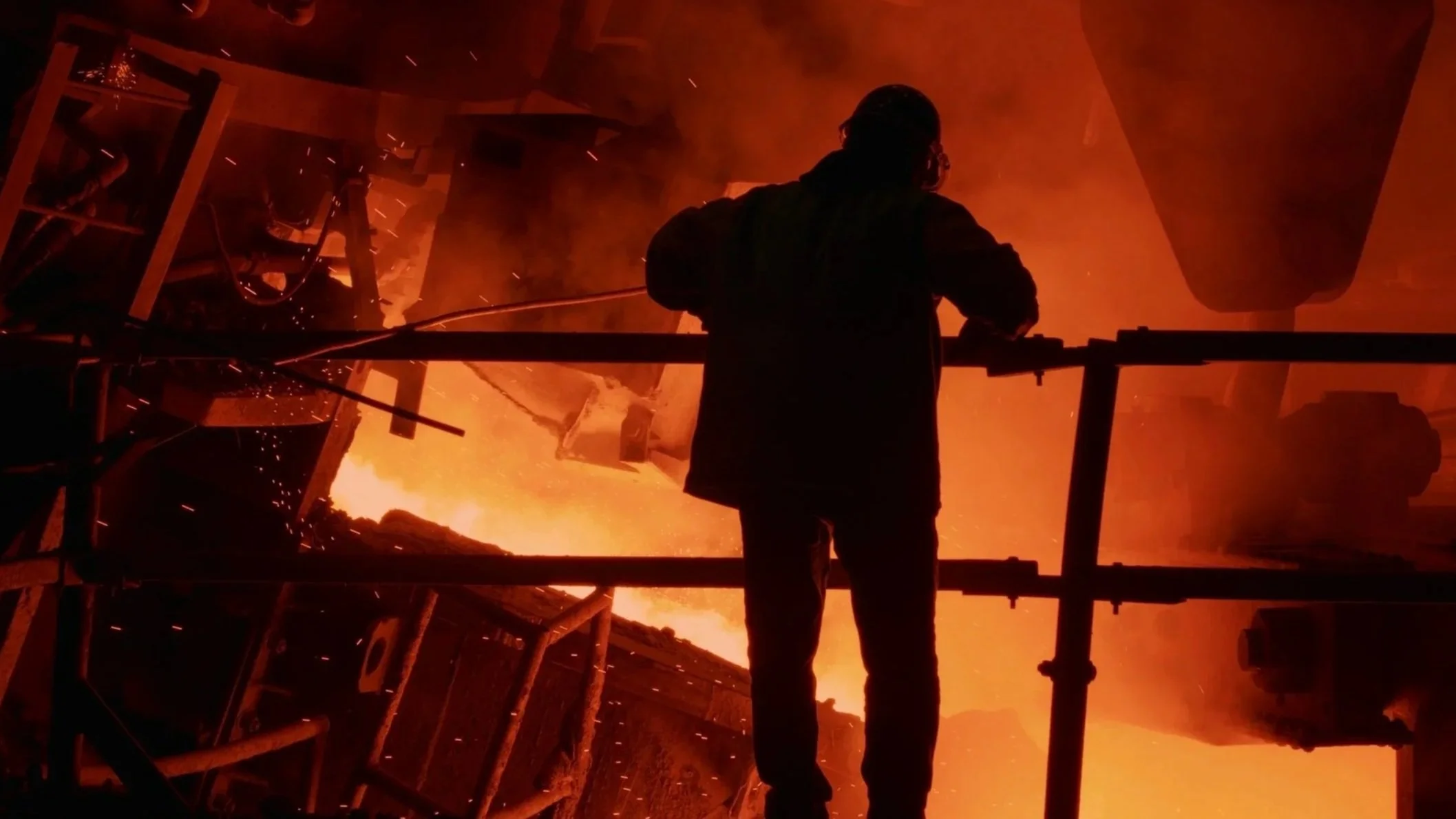 A worker stands on scaffolding inside a foundry furnace with bright orange flames and sparks in the background.