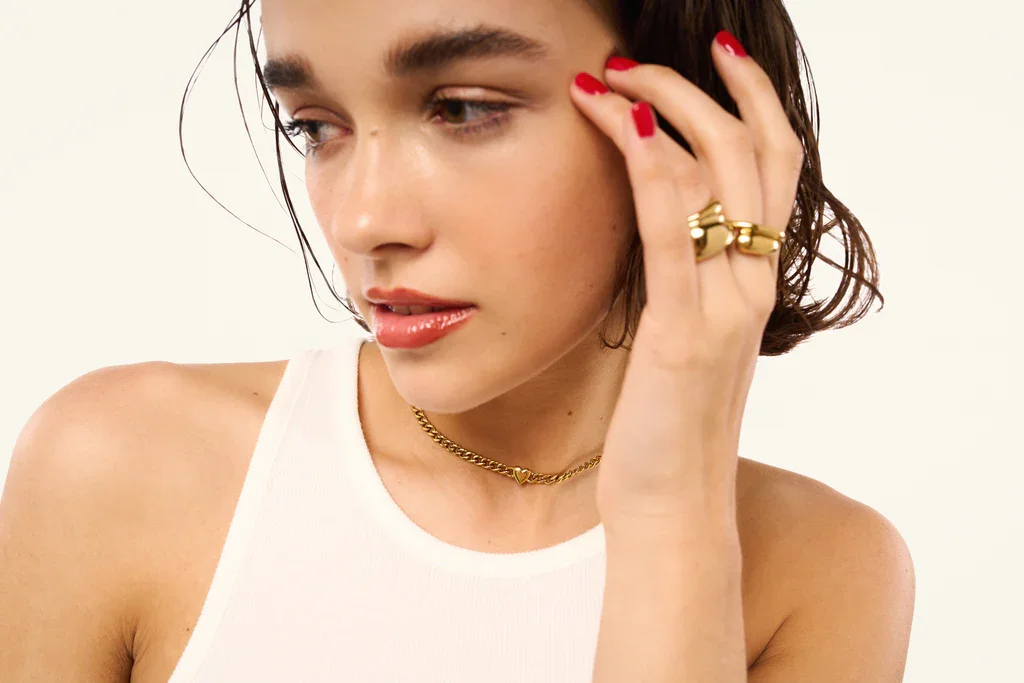A woman with blonde hair and green eyes wearing gold jewelry, including earrings, a chain bracelet, a ring, and a necklace, touching her face with her right hand.