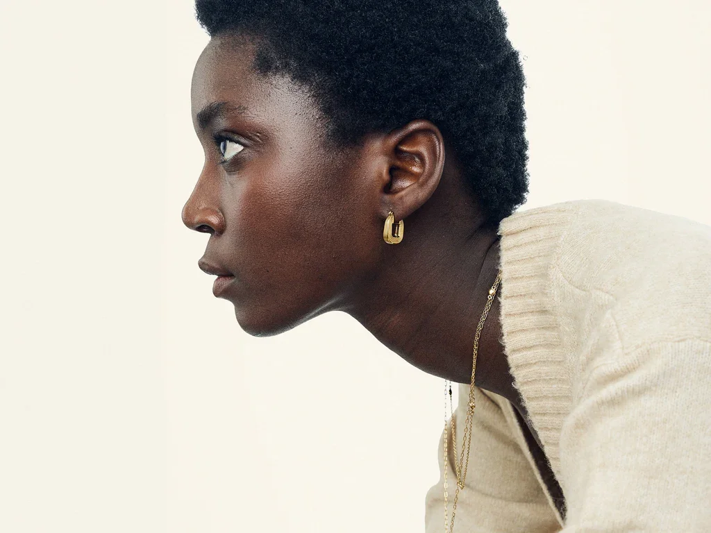 Close-up of a woman's neck and shoulder, wearing gold jewelry including a chain necklace and hoop earrings.