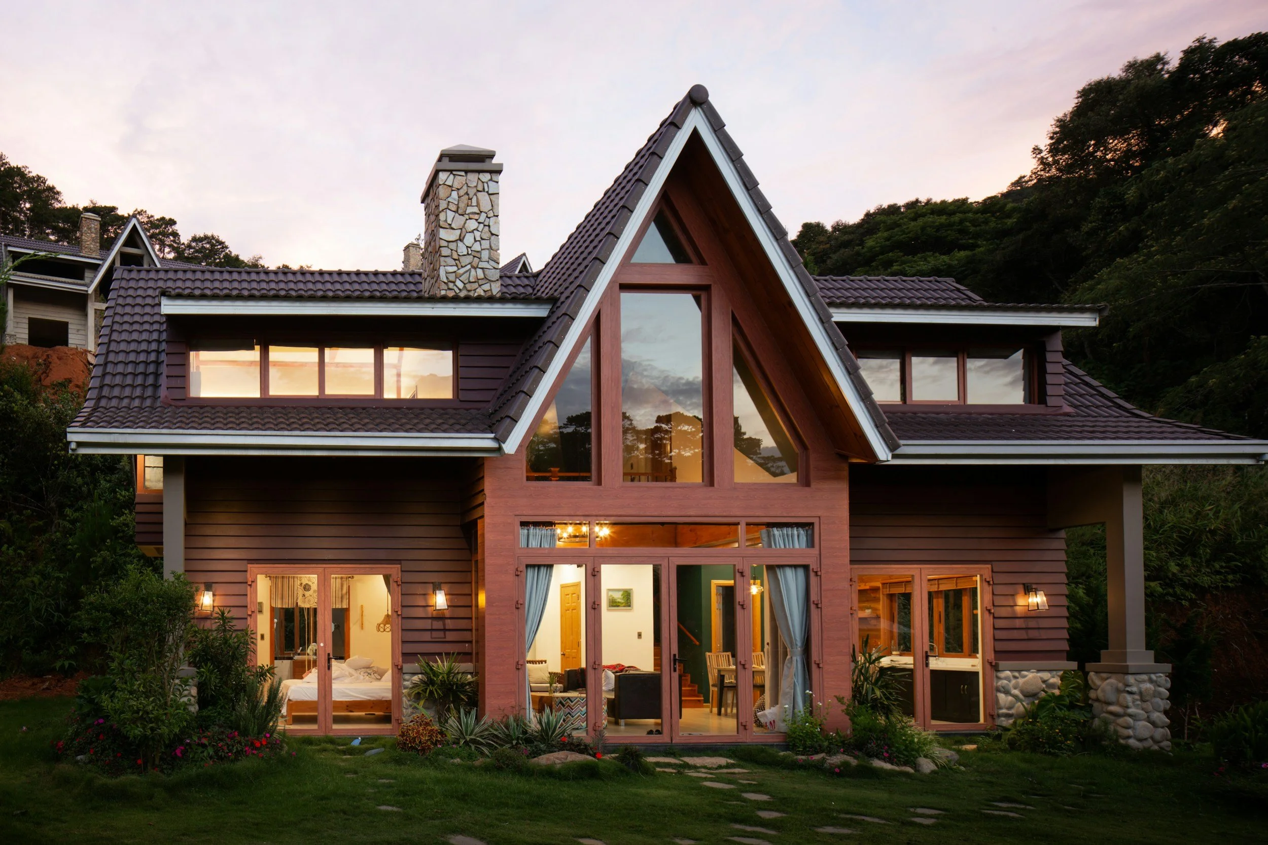 A house with wooden siding and large windows, illuminated from inside, set against a backdrop of trees and a colorful evening sky.