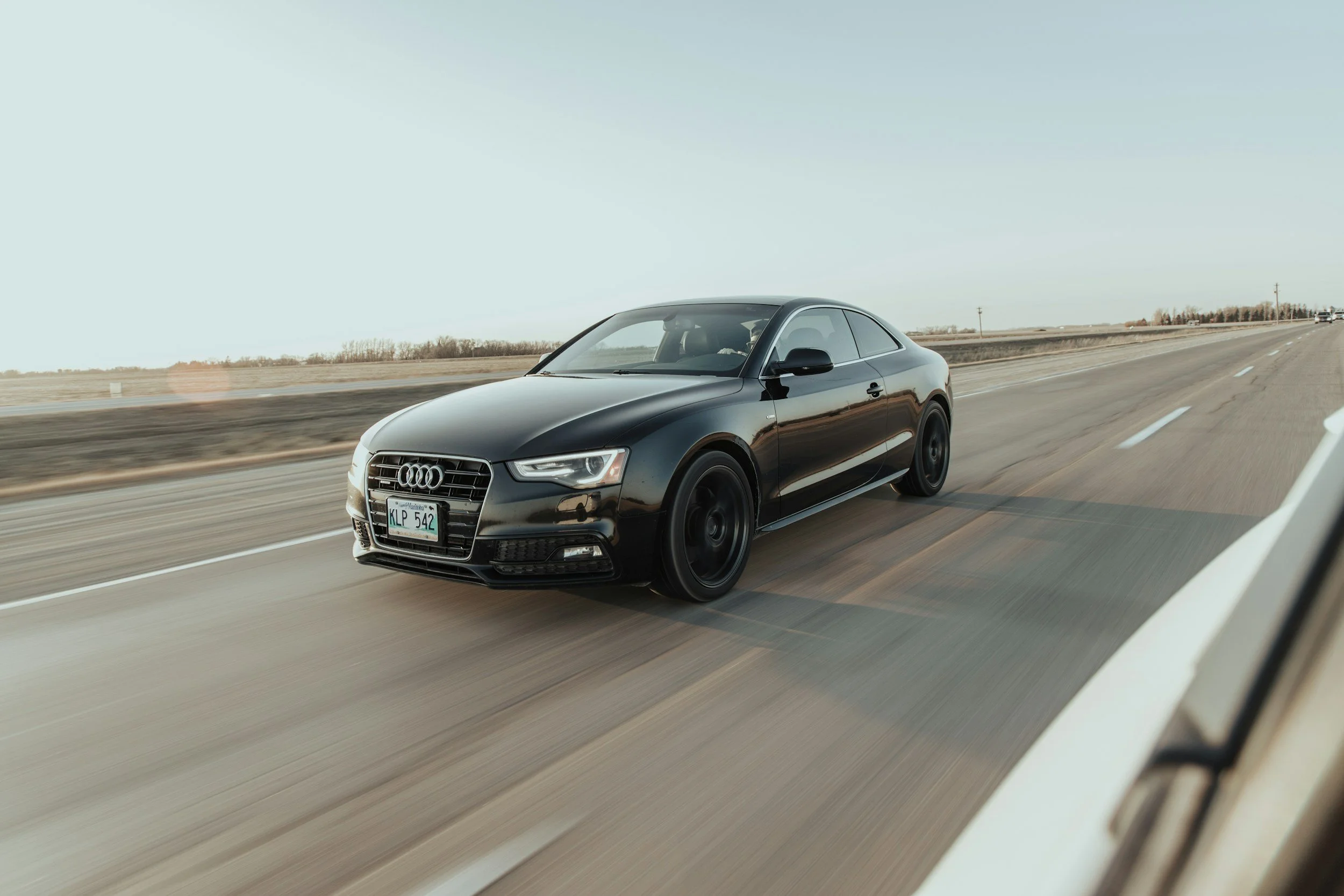 A black Audi coupe driving on an open highway with a clear sky.