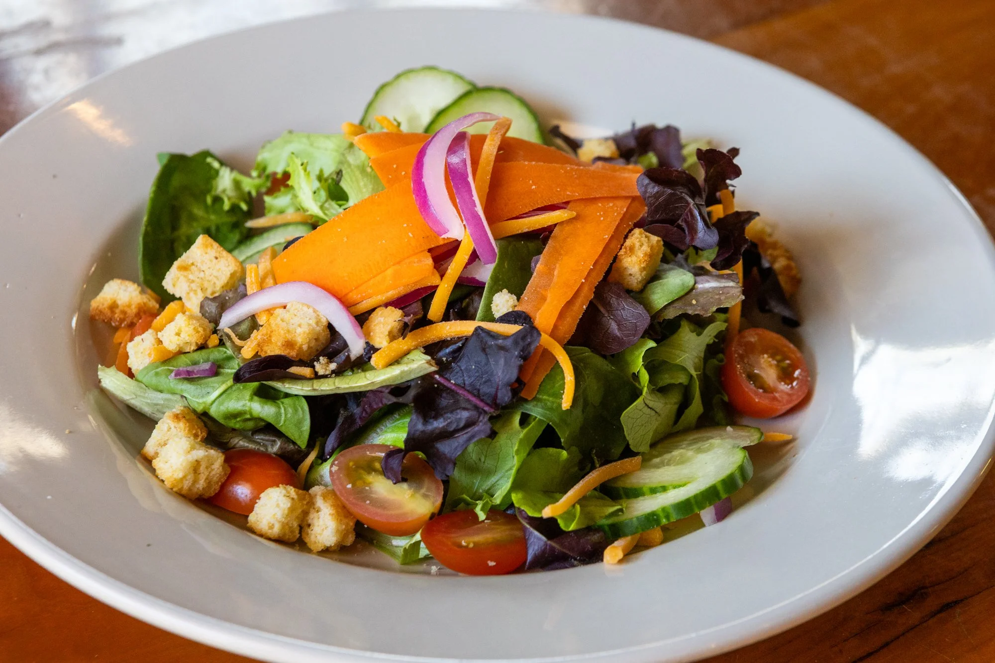 Fresh vegetable salad with cherry tomatoes, cucumbers, carrots, lettuce, red onions, shredded cheese, and croutons on a white plate.