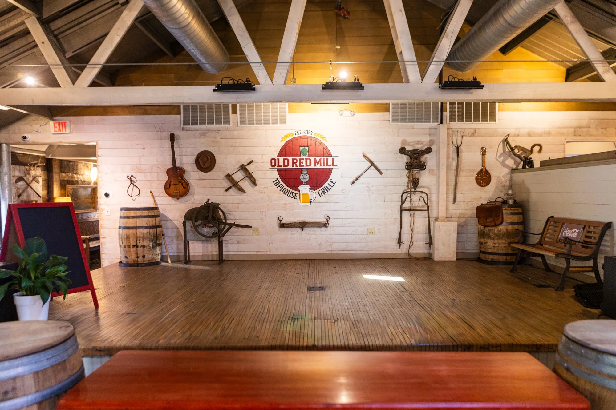 Empty rustic indoor stage area at Old Red Mill Taphouse Grill, decorated with vintage musical instruments, farm tools, and barrels, with a bench on the right and potted plant on the left.