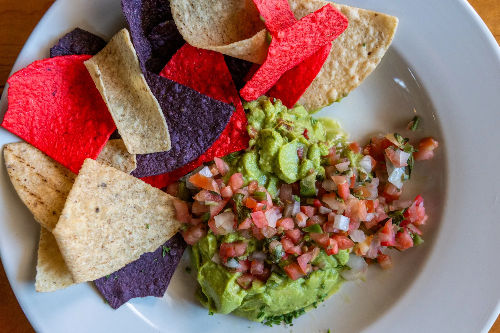 A white plate containing colorful tortilla chips, guacamole topped with pico de gallo, and sliced avocado.