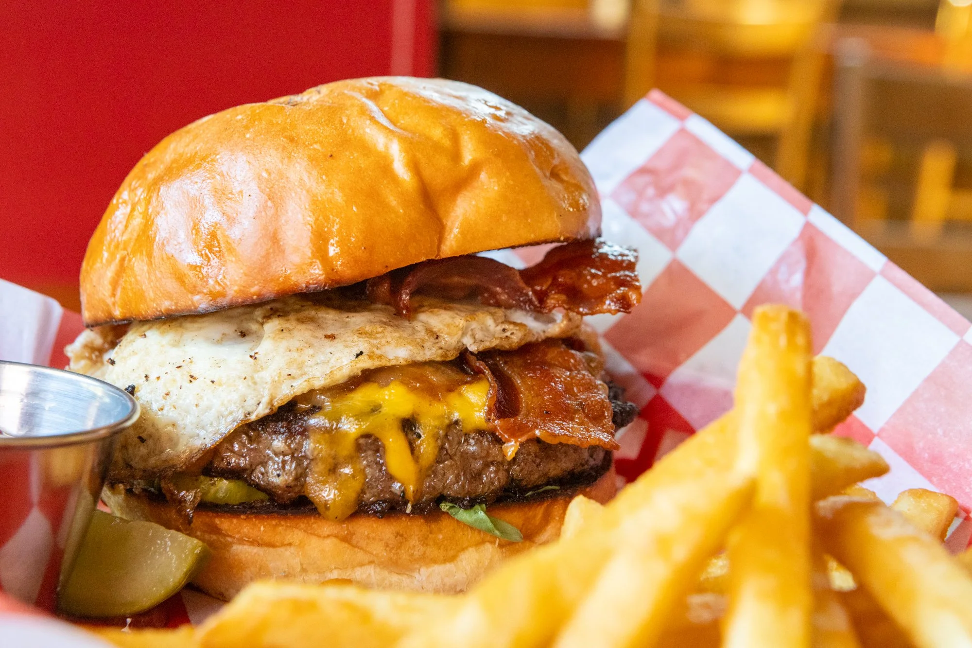 Close-up of a cheeseburger with bacon, fried egg, and melted cheese in a bun, served with French fries and pickles on a red checkered paper-lined basket.