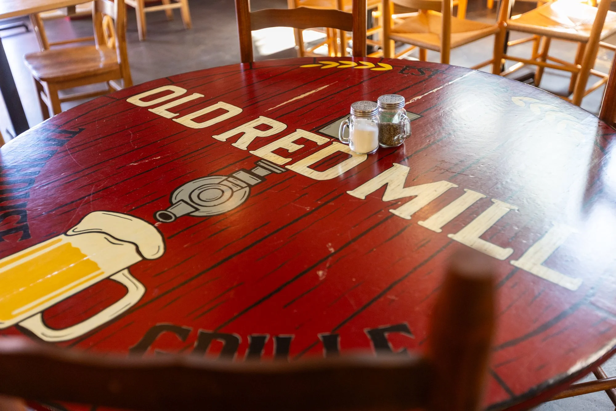 A round red wooden table with a printed logo and text, surrounded by wooden chairs in a restaurant. On the table are salt, pepper, and sugar shakers.