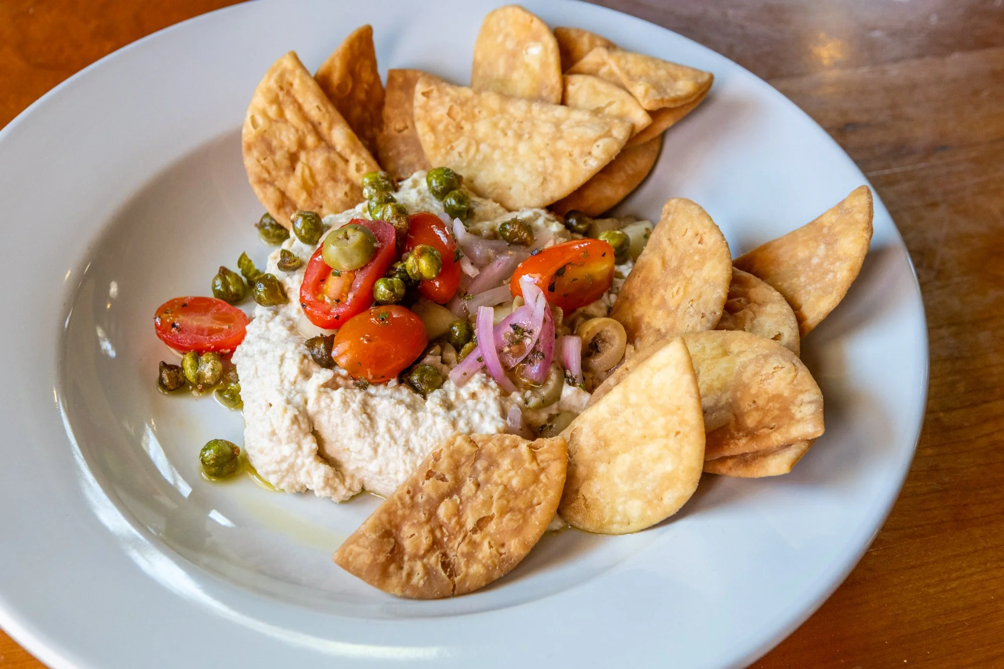 A plate of hummus topped with cherry tomatoes, green peas, and thinly sliced red onions, served with fried pita chips.