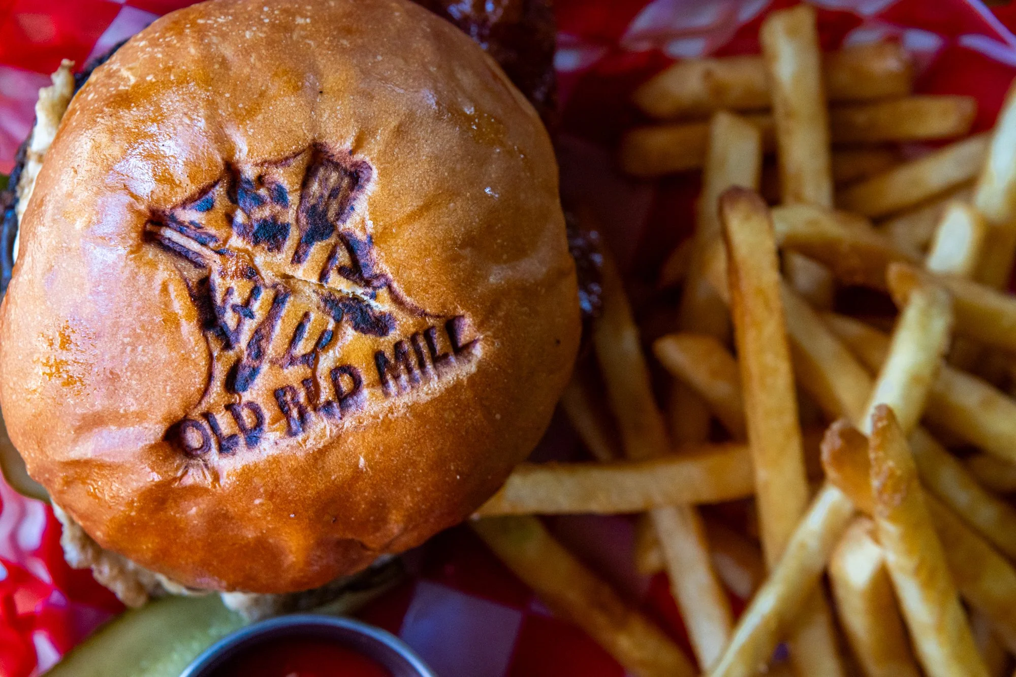 A burger with a stamped bun featuring the Olde Bradford Mill logo, accompanied by French fries and a small container of ketchup.