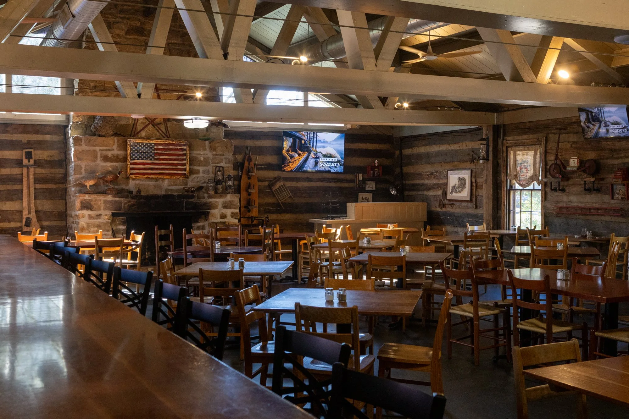 Empty rustic restaurant or barn-style dining area with wooden tables and chairs, American flag artwork on stone fireplace, mounted TVs, and vintage decorations, illuminated by natural and artificial light.
