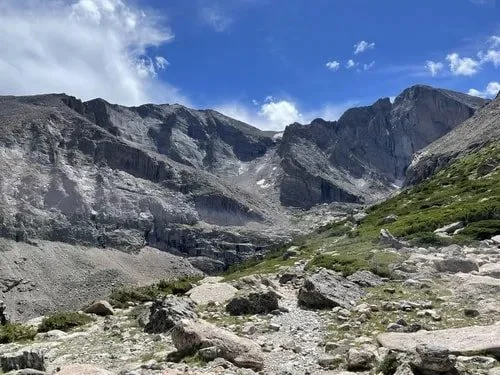 Mountain landscape with rocky terrain, sparse green vegetation, and a partly cloudy blue sky.