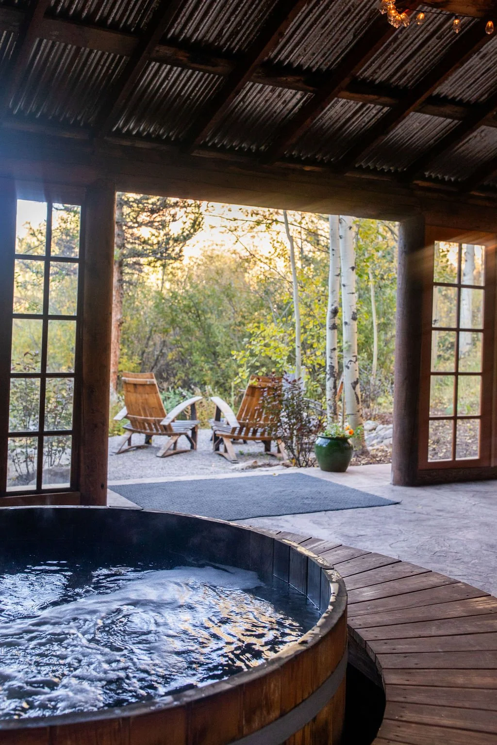 View from inside a rustic cabin with a hot tub, looking out onto a patio with two wooden chairs and lush trees in the background during sunset.