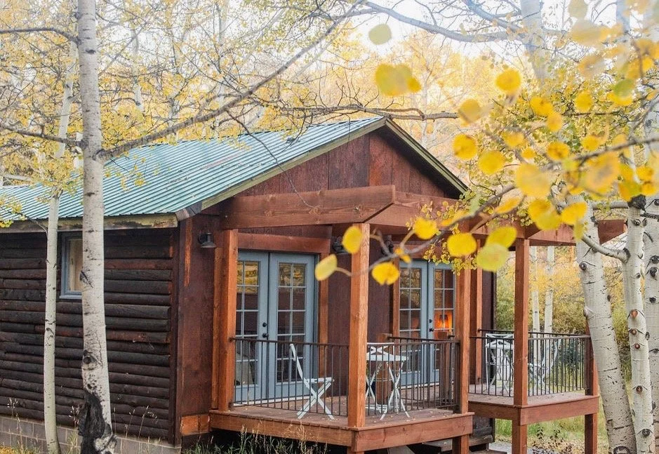 A cozy cabin with a wooden deck and patio furniture, surrounded by autumn trees with yellow leaves.