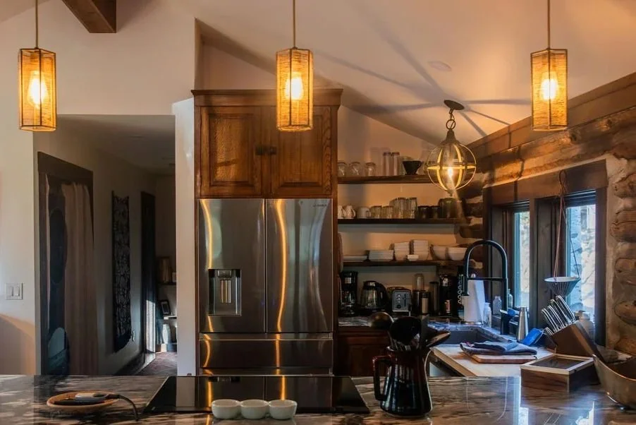 Kitchen with wooden cabinetry, stainless steel refrigerator, open shelves with dishes and mugs, black faucet, window with view outside, hanging pendant lights, and stone wall on the right.