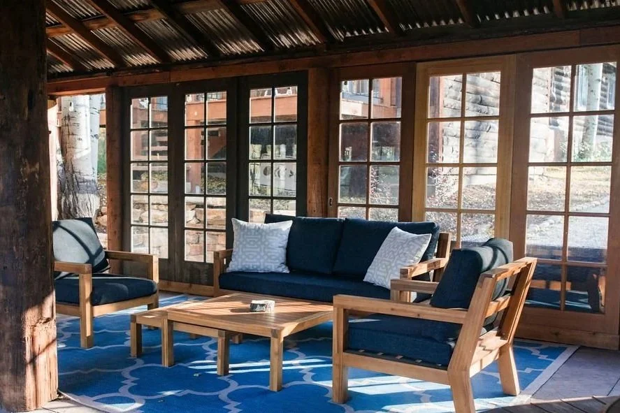 Sunlit porch with wooden furniture, including a navy blue sofa and matching chairs with white pillows, a wooden coffee table, a blue patterned rug, large windows with wooden frames, and trees visible outside.