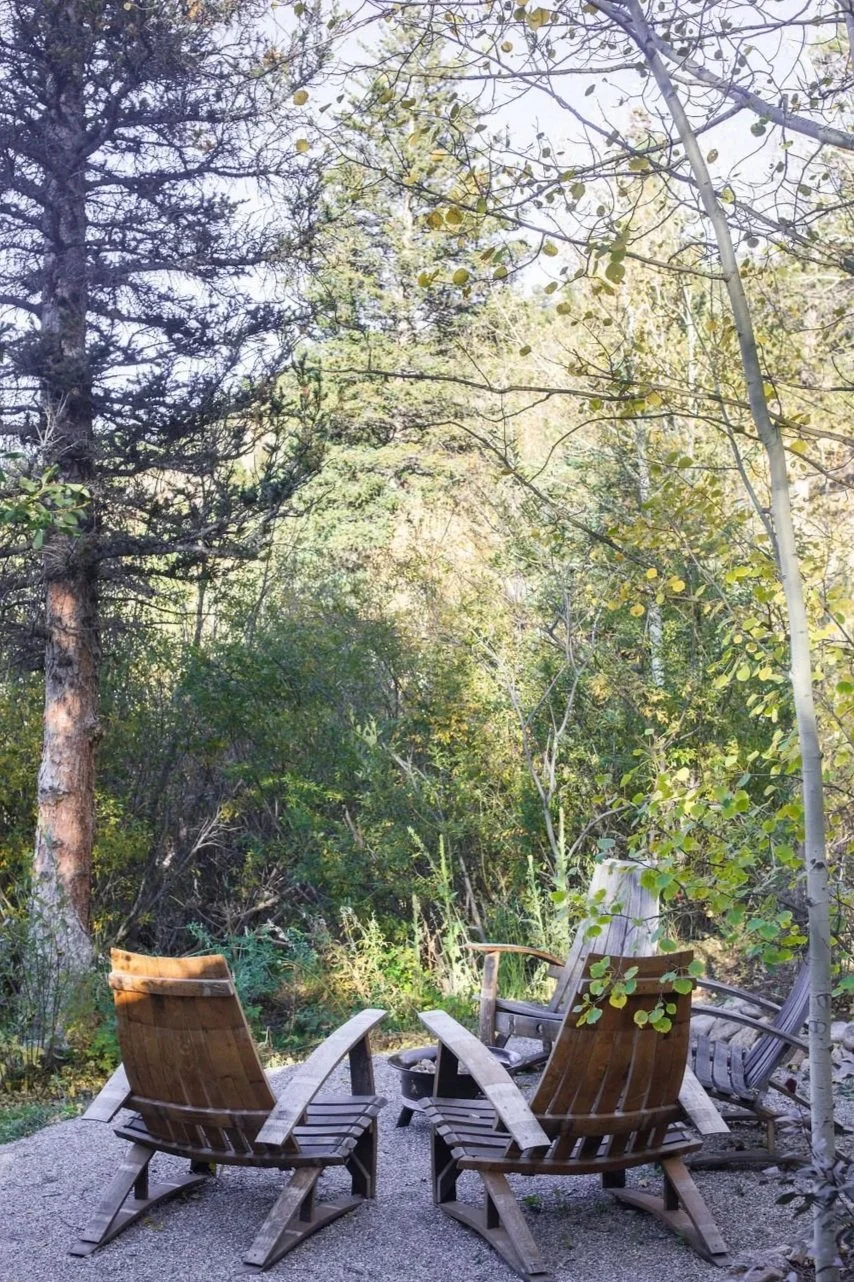 Three wooden chairs around a fire pit on a gravel surface in a lush, green forest with tall trees and foliage.
