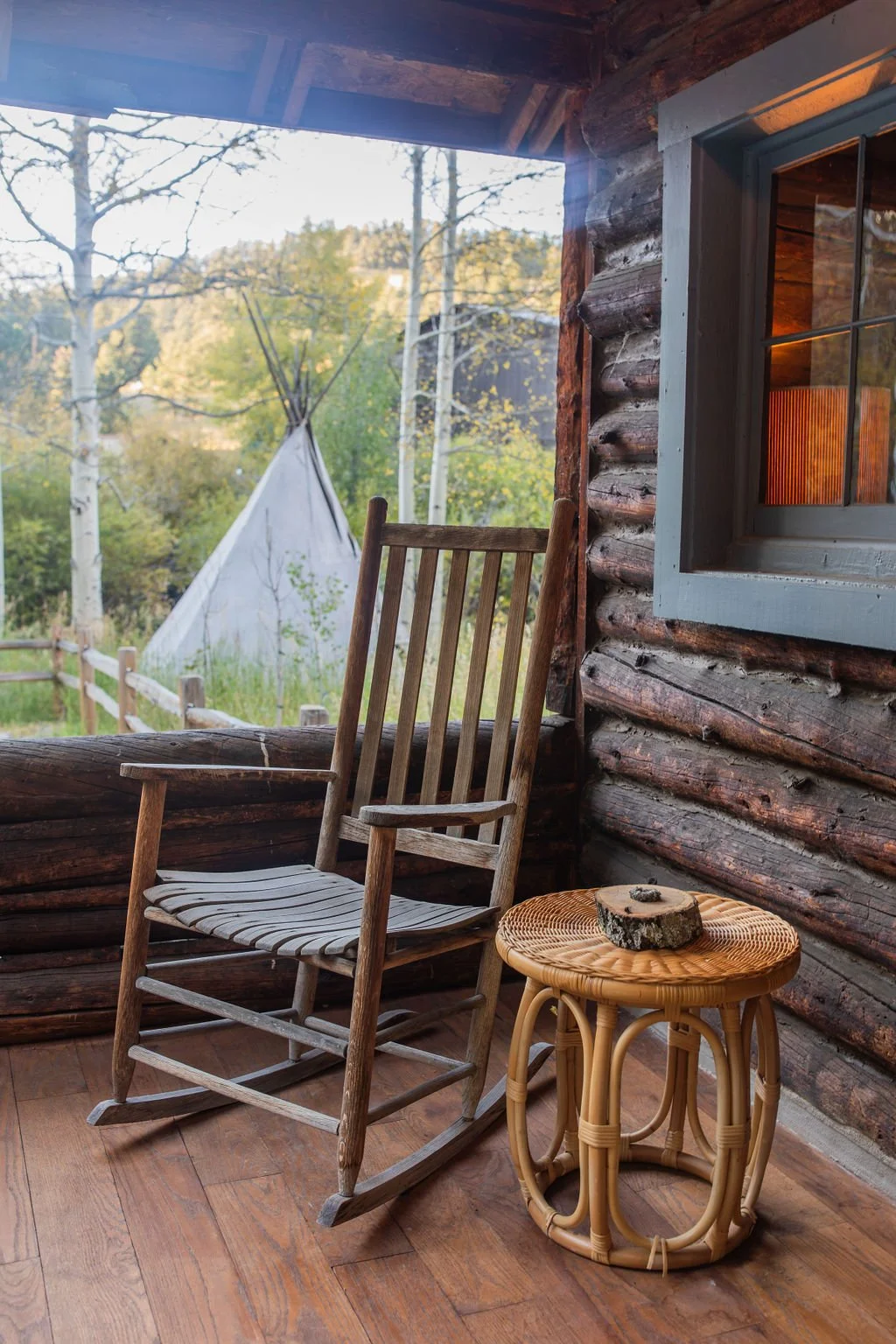 View from a rustic cabin porch showing a wooden rocking chair next to a small wicker table with a piece of wood on top, and a teepee in a wooded outdoor setting.