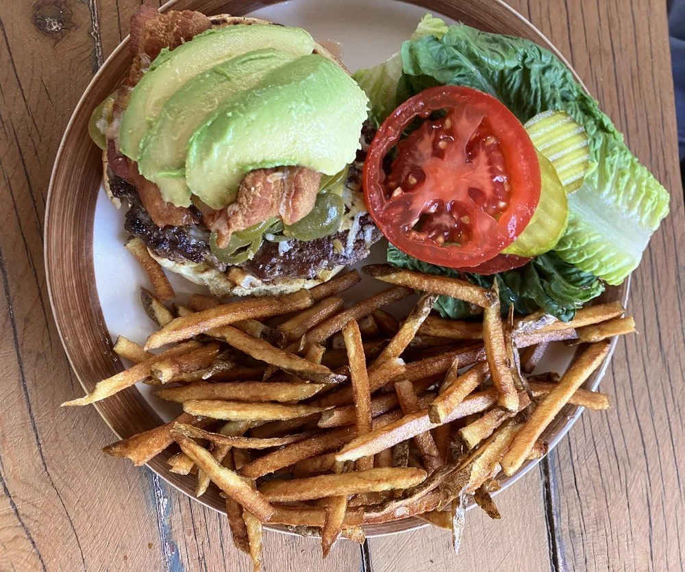 Burgers with sliced avocado, lettuce, tomato, pickles, and French fries on a plate.