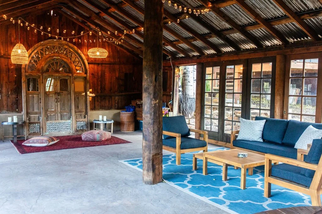 A cozy living space with wooden walls and ceiling, string lights, and large windows. The room features a navy blue sofa with white pillows, a matching armchair, a wooden coffee table, and a blue patterned rug. There are also cushions and a small red rug with pillows on the floor, creating a relaxed atmosphere.