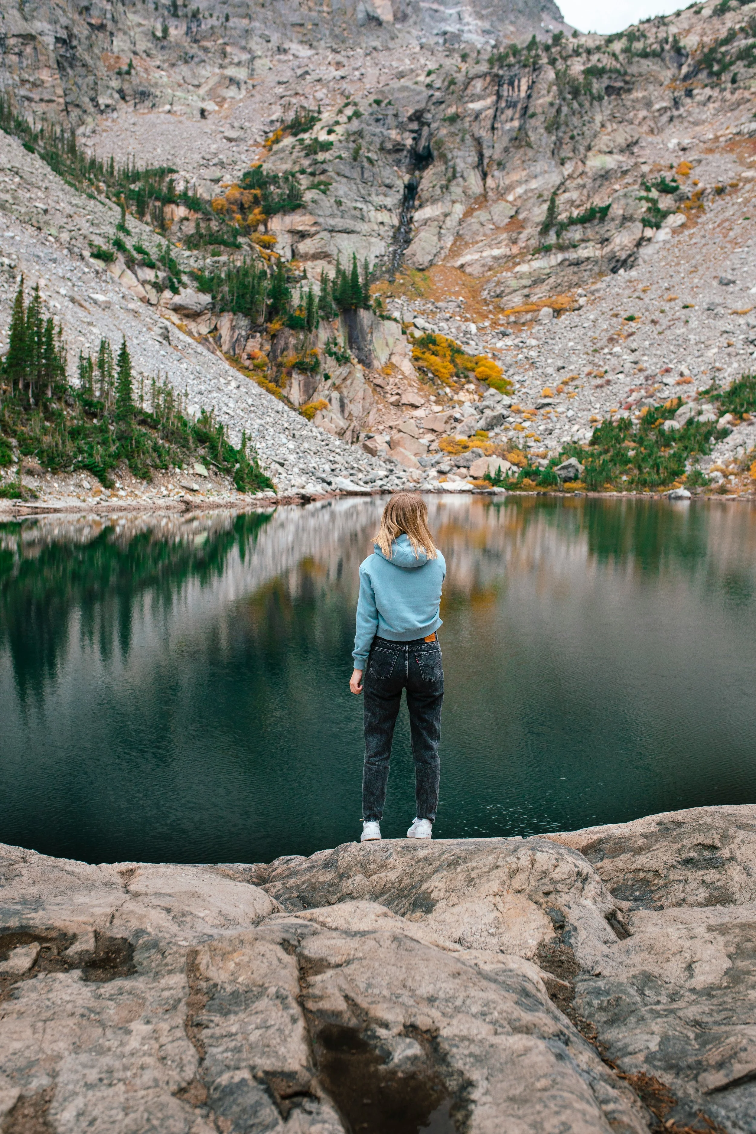 A woman standing on rocks near a calm lake, surrounded by mountains with autumn foliage.