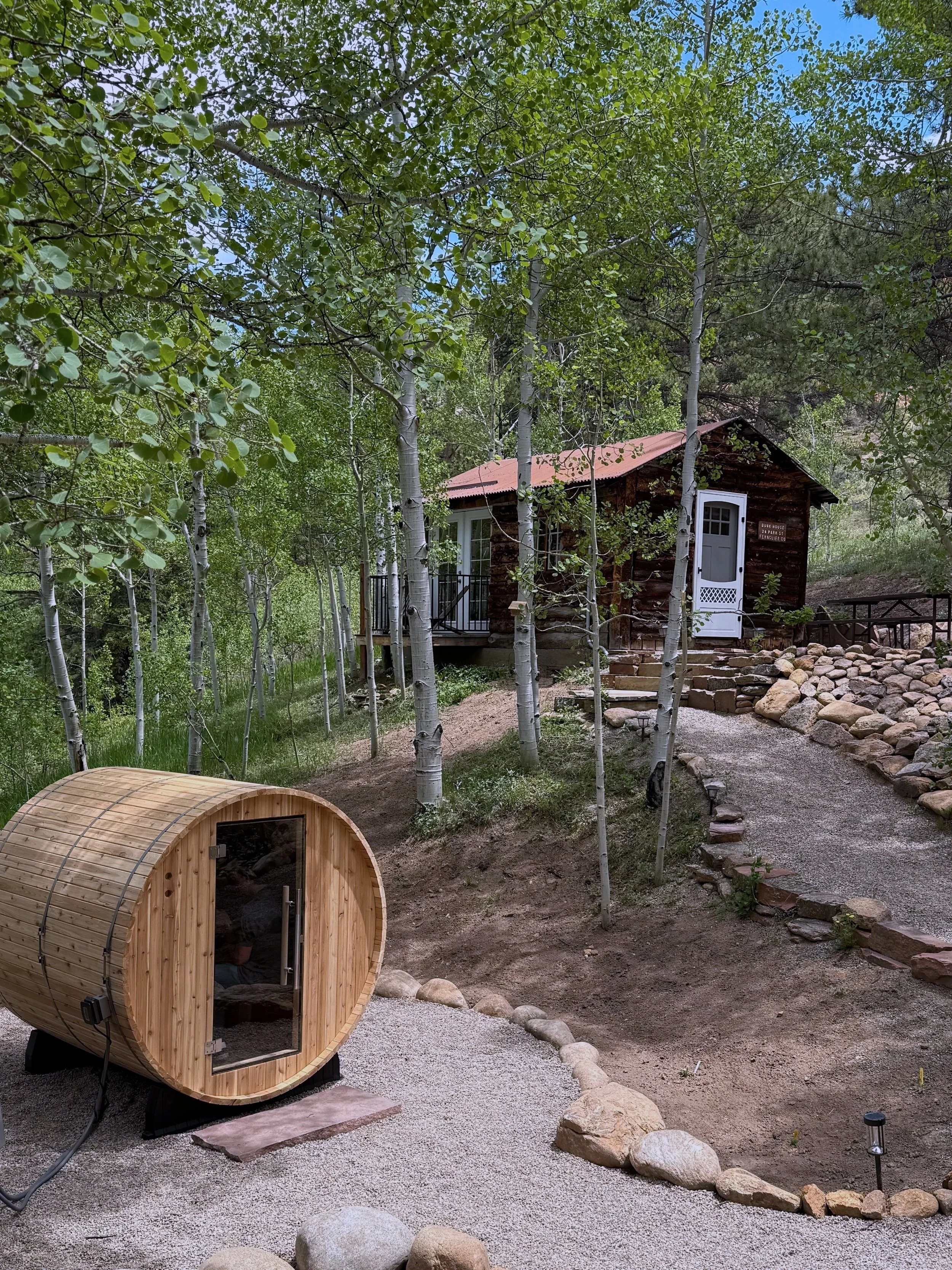 A small wooden cabin with a red metal roof surrounded by trees, with a gravel pathway and a wood sauna in the foreground.