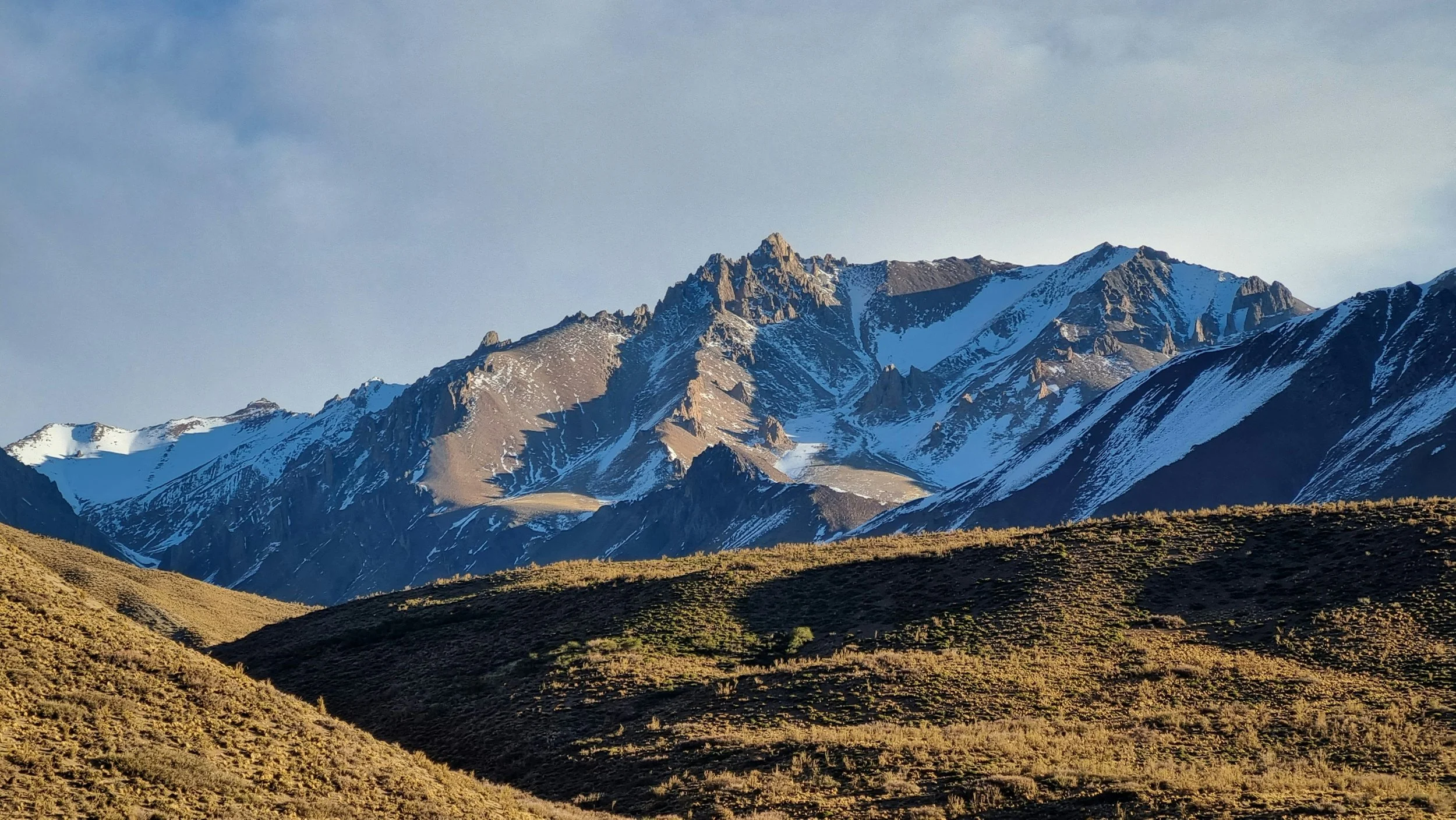 Mountain range with snow-capped peaks and rolling grassy hills in the foreground.