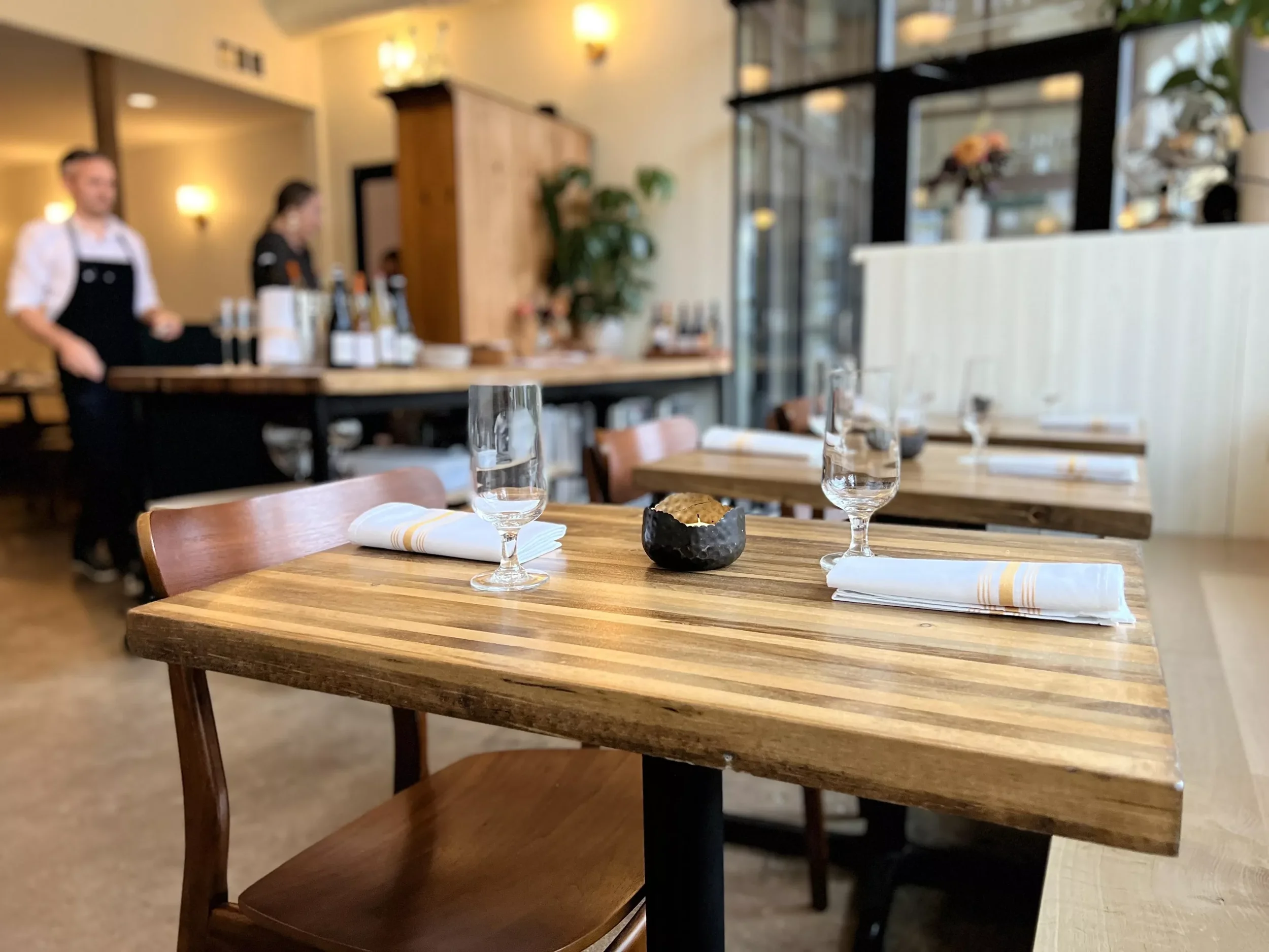 Restaurant table set with two water glasses, napkins, and a small bowl, with blurred background of restaurant interior and staff.