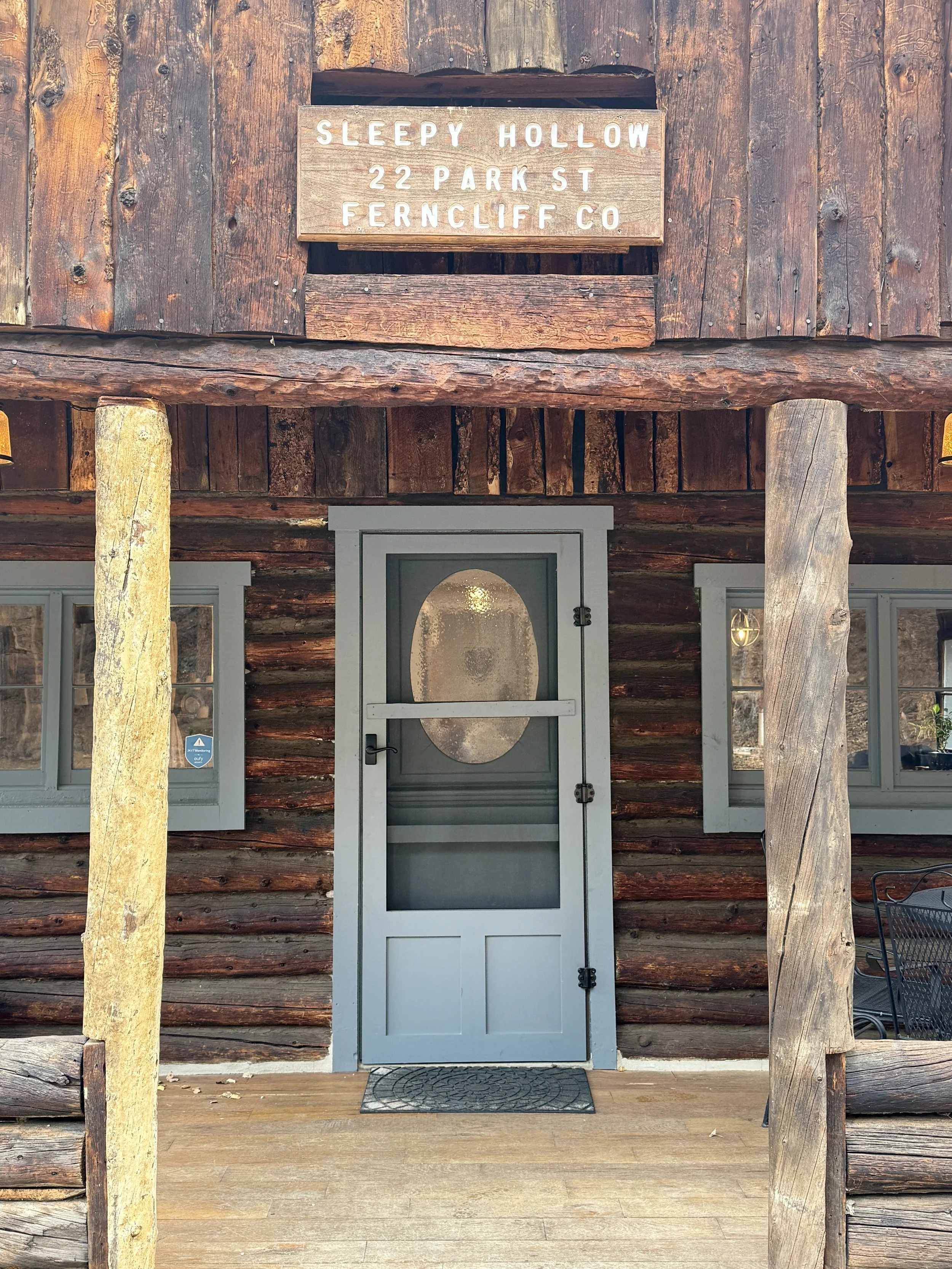 Front entrance of a rustic log cabin with a blue door and a sign above it reading 'Sleepy Hollow, 22 Park St, Ferncliff Co'.
