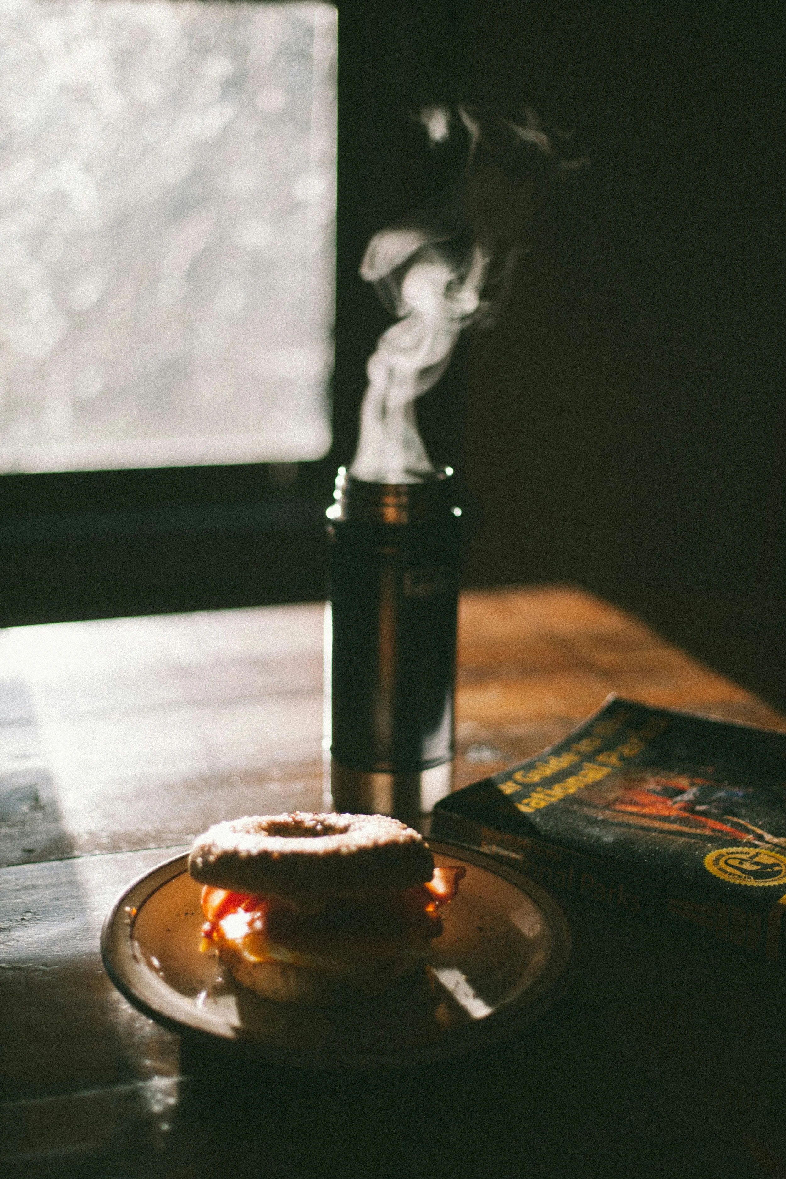 A vape pen emitting vapor, a partially eaten donut on a plate with caramel sauce, a book titled 'Essential Oils' on a wooden surface, and a computer monitor in the background.