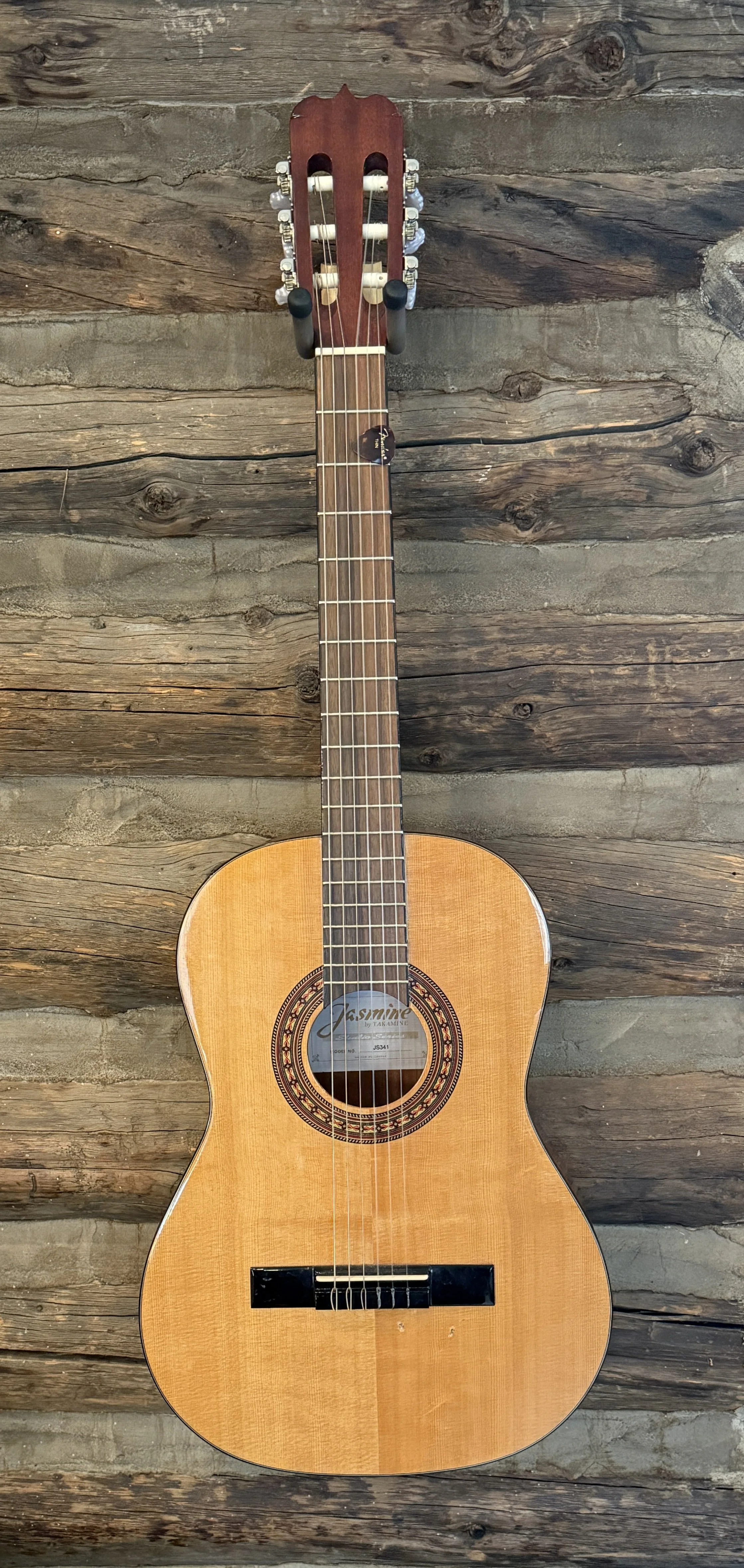 An acoustic guitar with a wooden body, six strings, and a dark wooden neck, mounted against a rustic wooden wall.