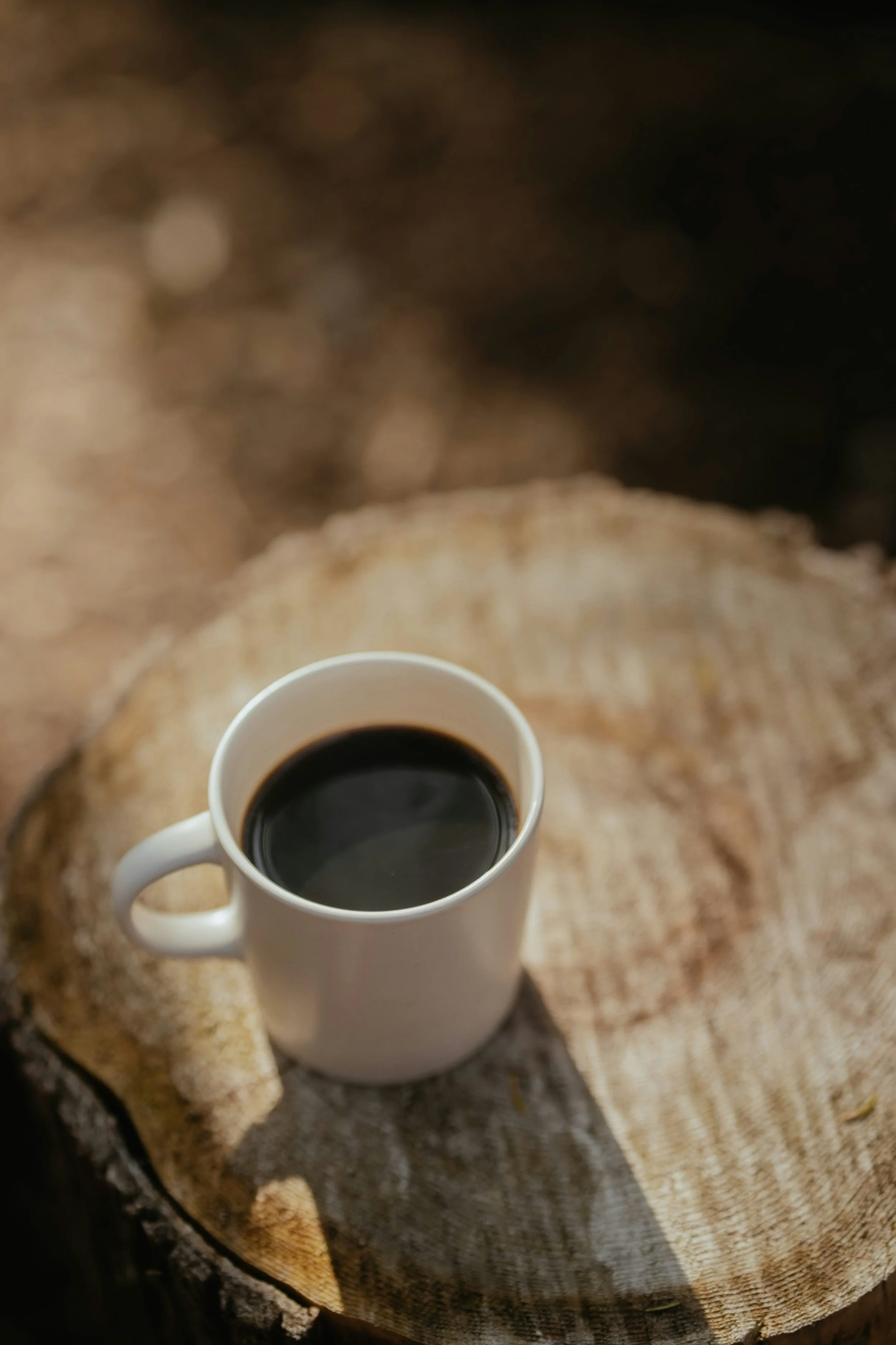 A white mug filled with black coffee placed on a cut tree trunk used as a table in a natural setting.