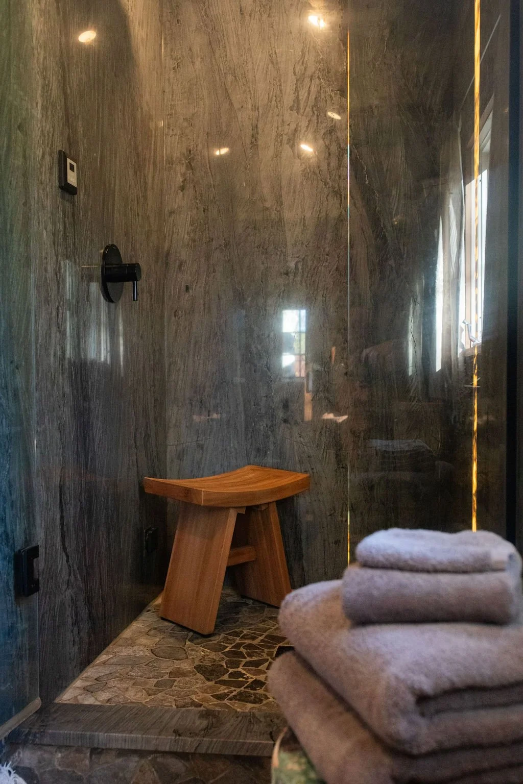 A steam room with dark stone walls, a small wooden bench, and a stone-tiled floor. Towels are folded on a surface in the foreground, and a window with natural light is reflected on the wall.