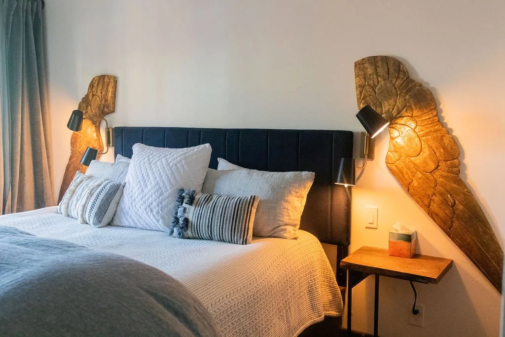 Bedroom with bed, pillows, wooden wall art, and side table with lamp.