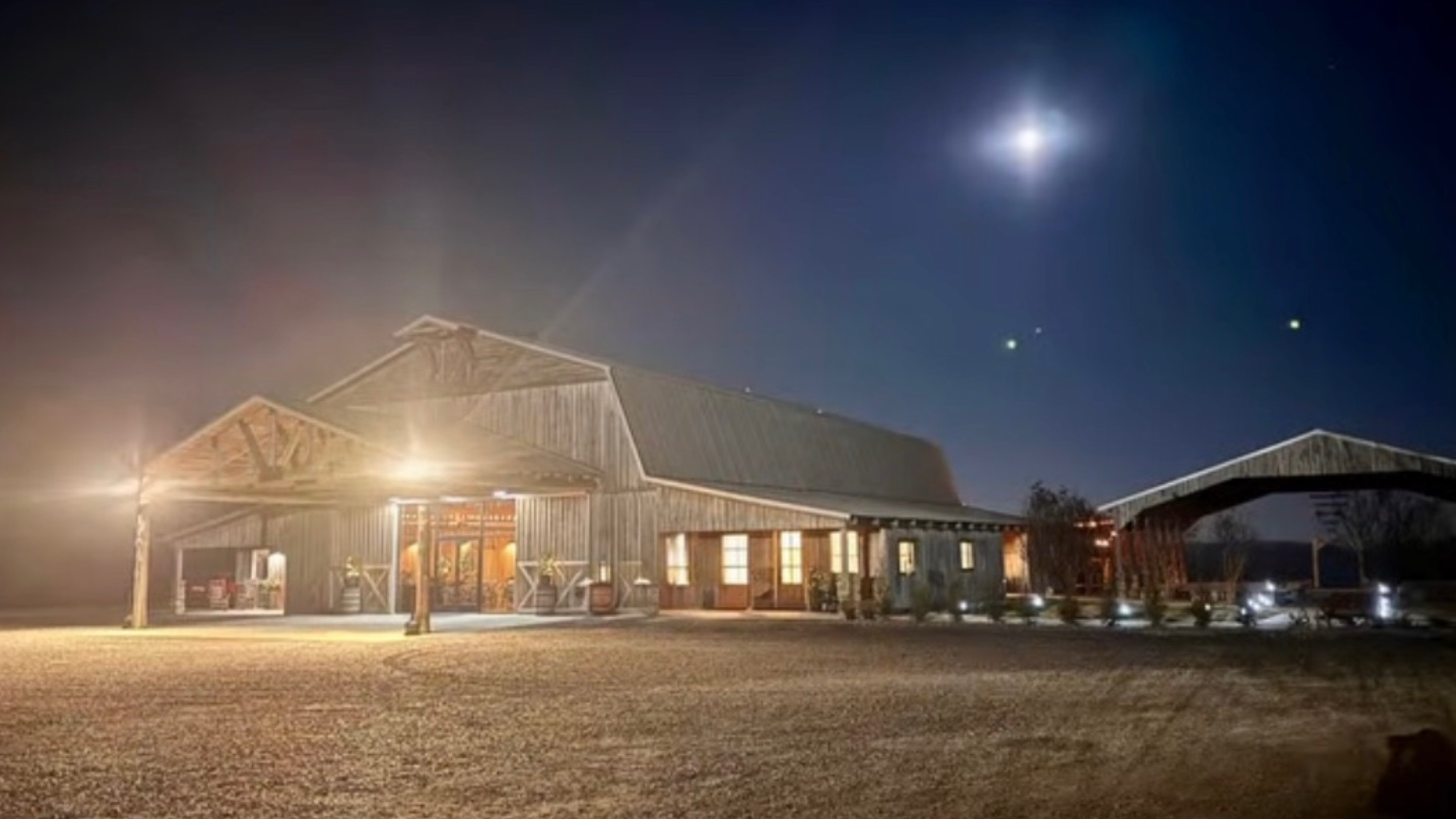 Nighttime view of a rustic barn with warm lights, a covered walkway, and a gravel ground under a starry sky with a bright moon and visible planets.