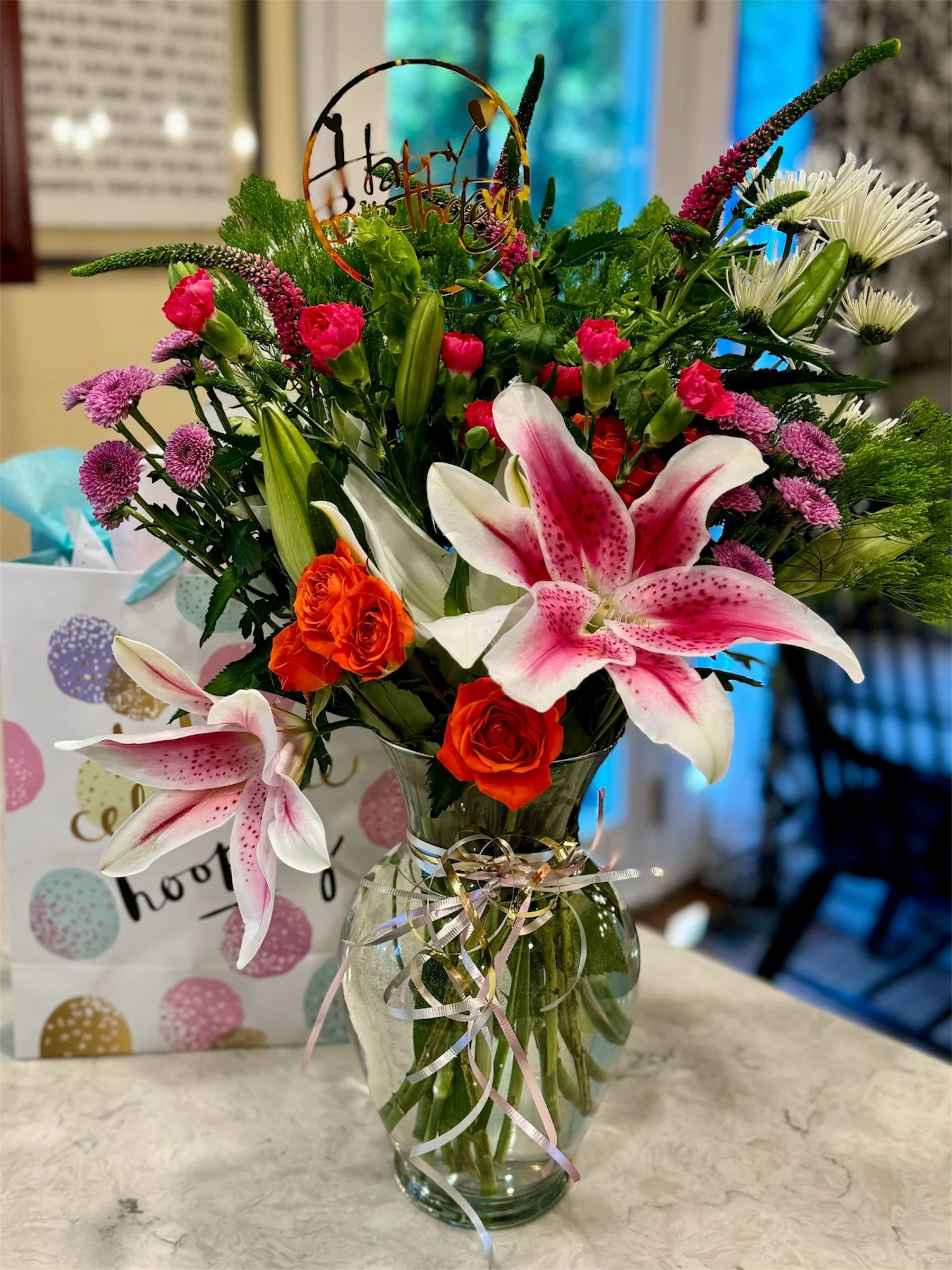 A vibrant bouquet of mixed flowers in a clear glass vase on a marble surface, with a white gift bag featuring colorful polka dots and the words "hooray" in the background.