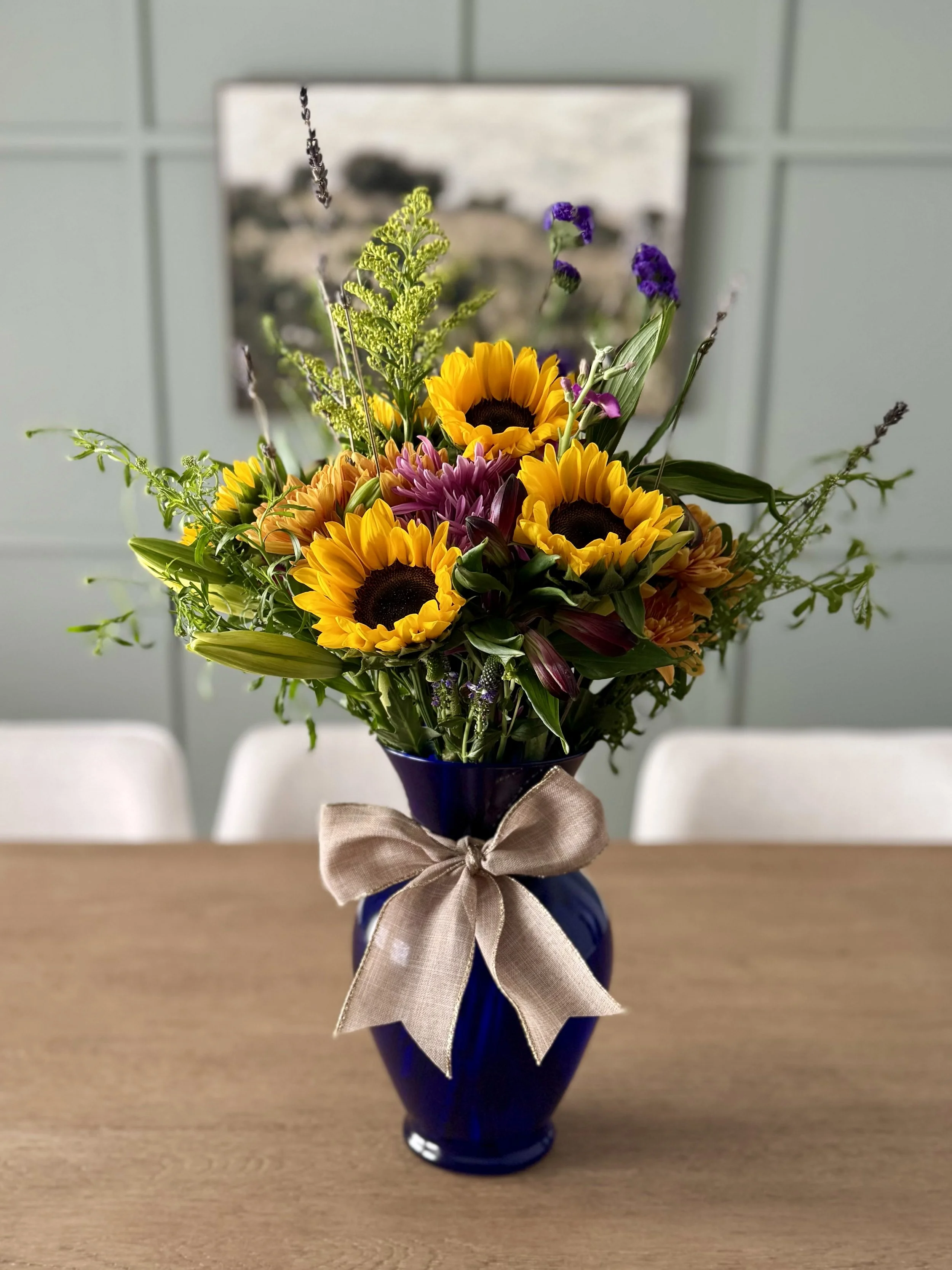 A bouquet of sunflowers, purple flowers, and greenery in a dark blue vase with a beige ribbon, placed on a wooden table.