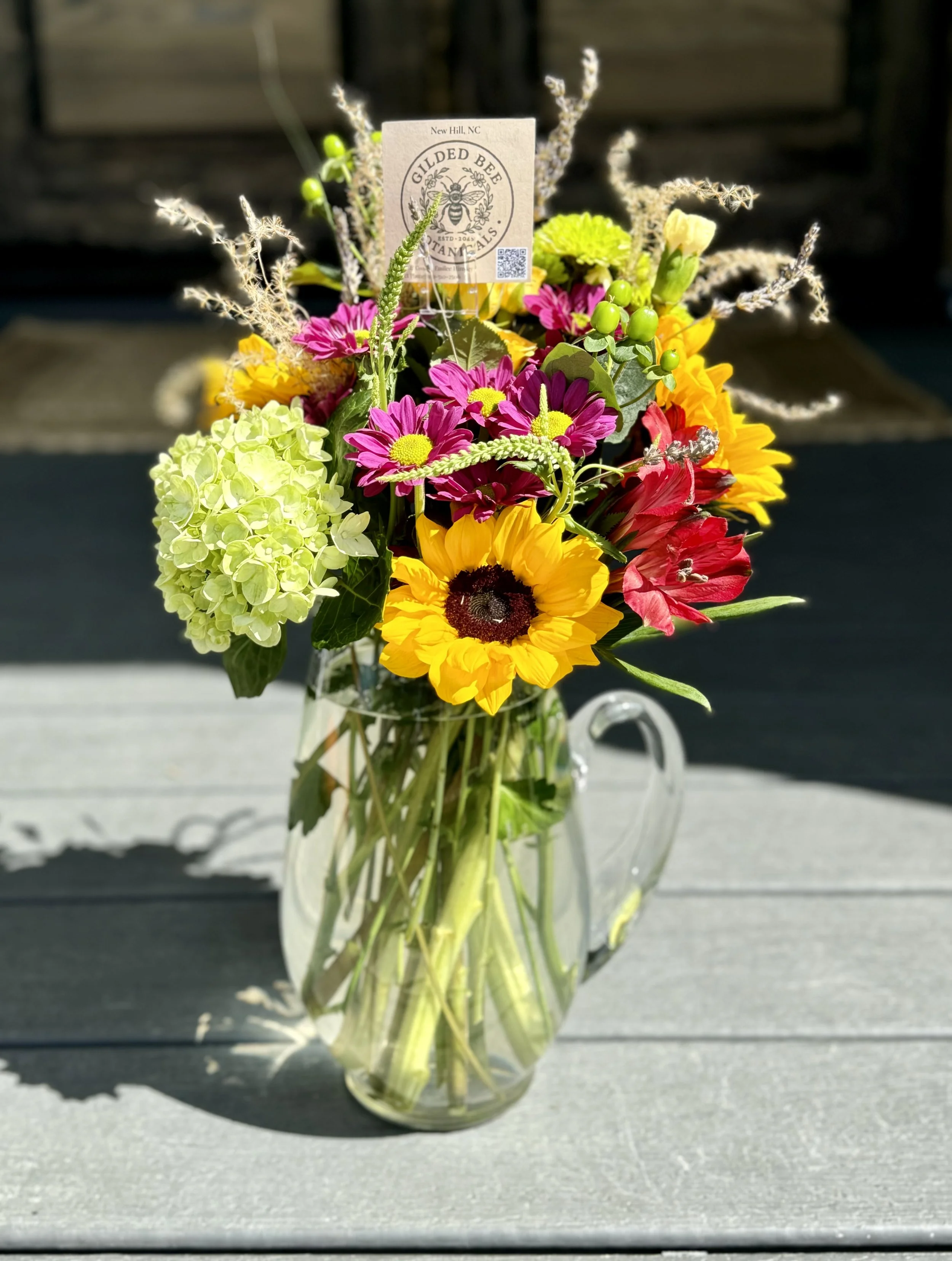 A colorful bouquet of flowers in a clear glass pitcher, including sunflowers, pink daisies, green hydrangeas, and other flowers, with a informational tag on some of the flowers.