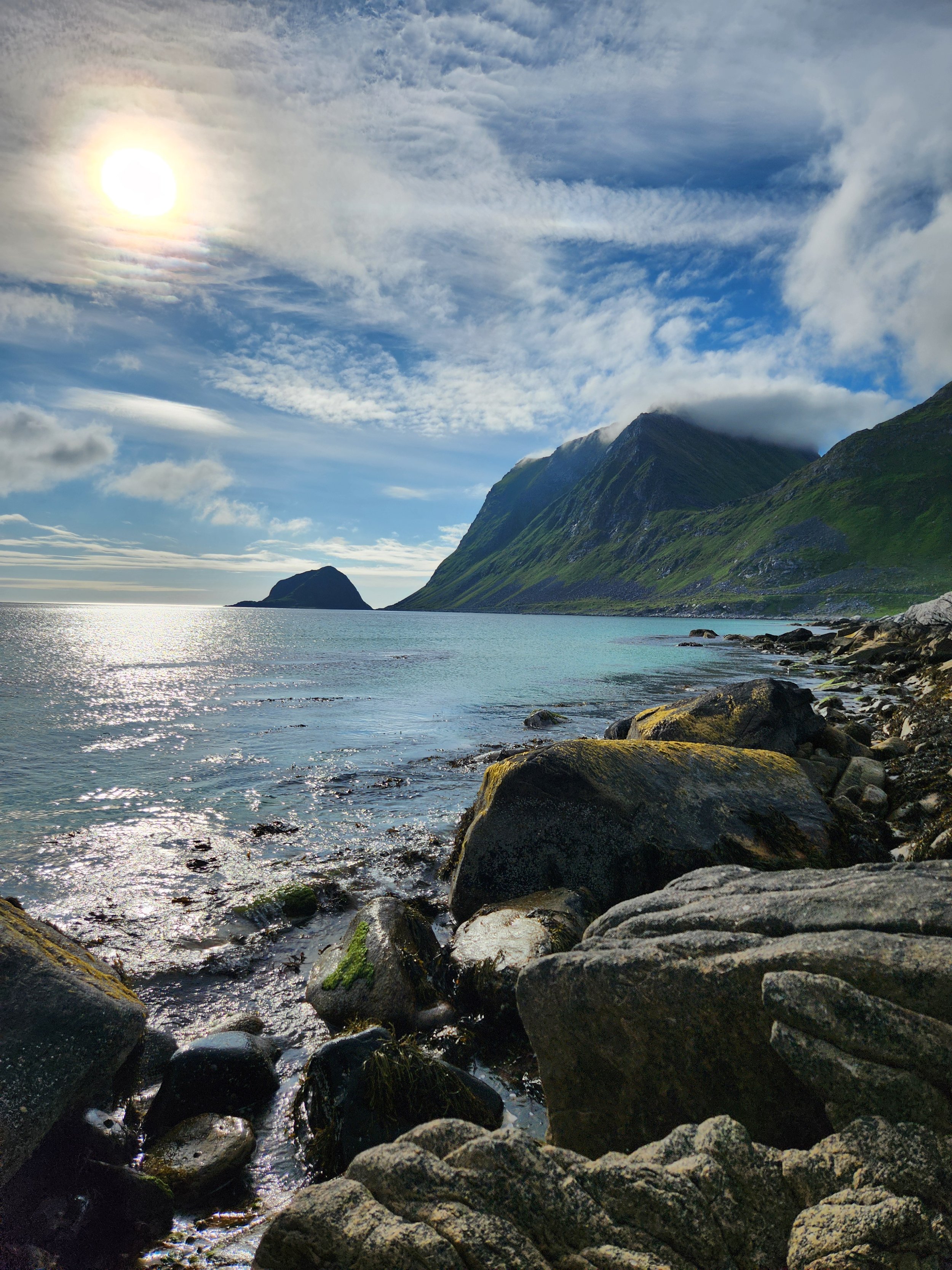 A scenic coastal landscape with rocky shoreline, calm water, green mountains, partly cloudy sky, and the sun shining.