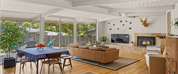 Living room with a dining table, a leather sofa, and a fireplace with a mounted TV, large windows, and decorative decor