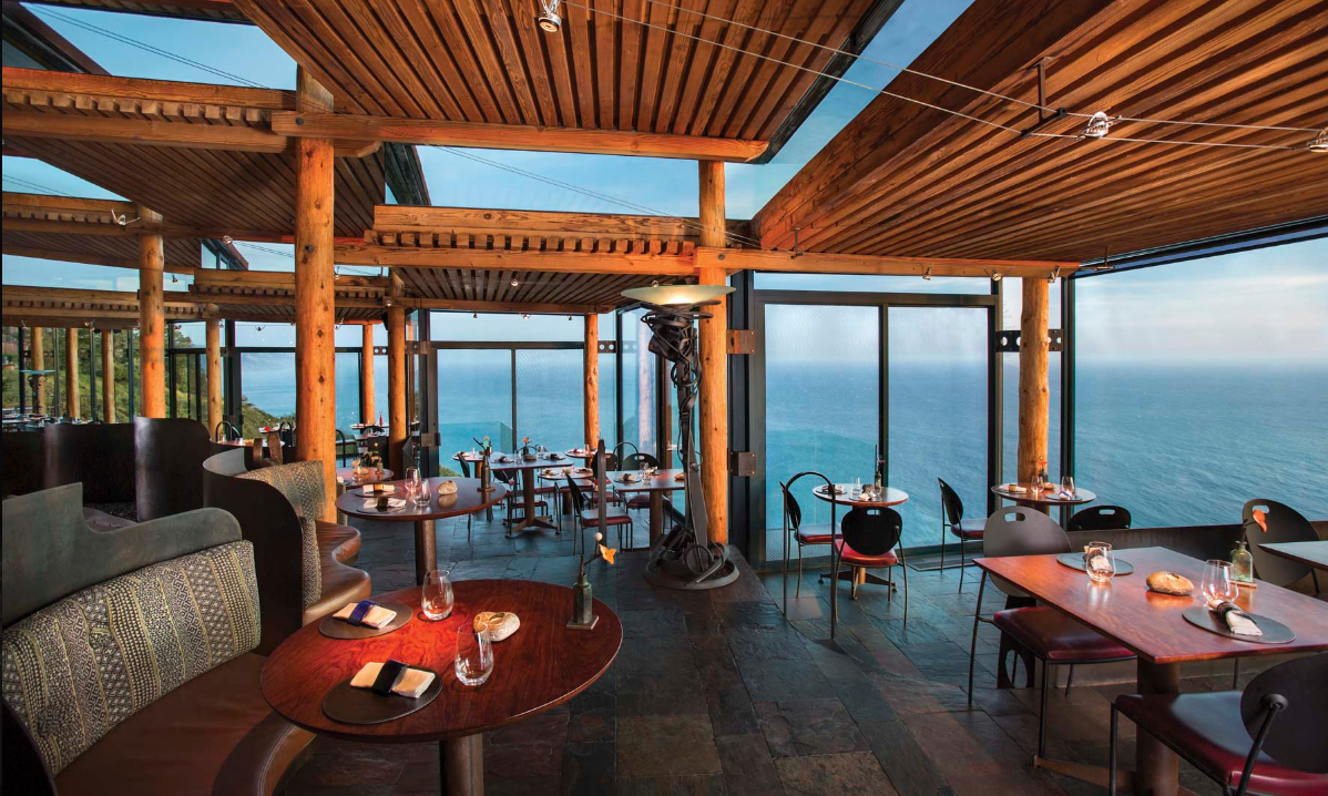 Interior of a restaurant with wooden ceiling and large glass windows overlooking the ocean during the day.