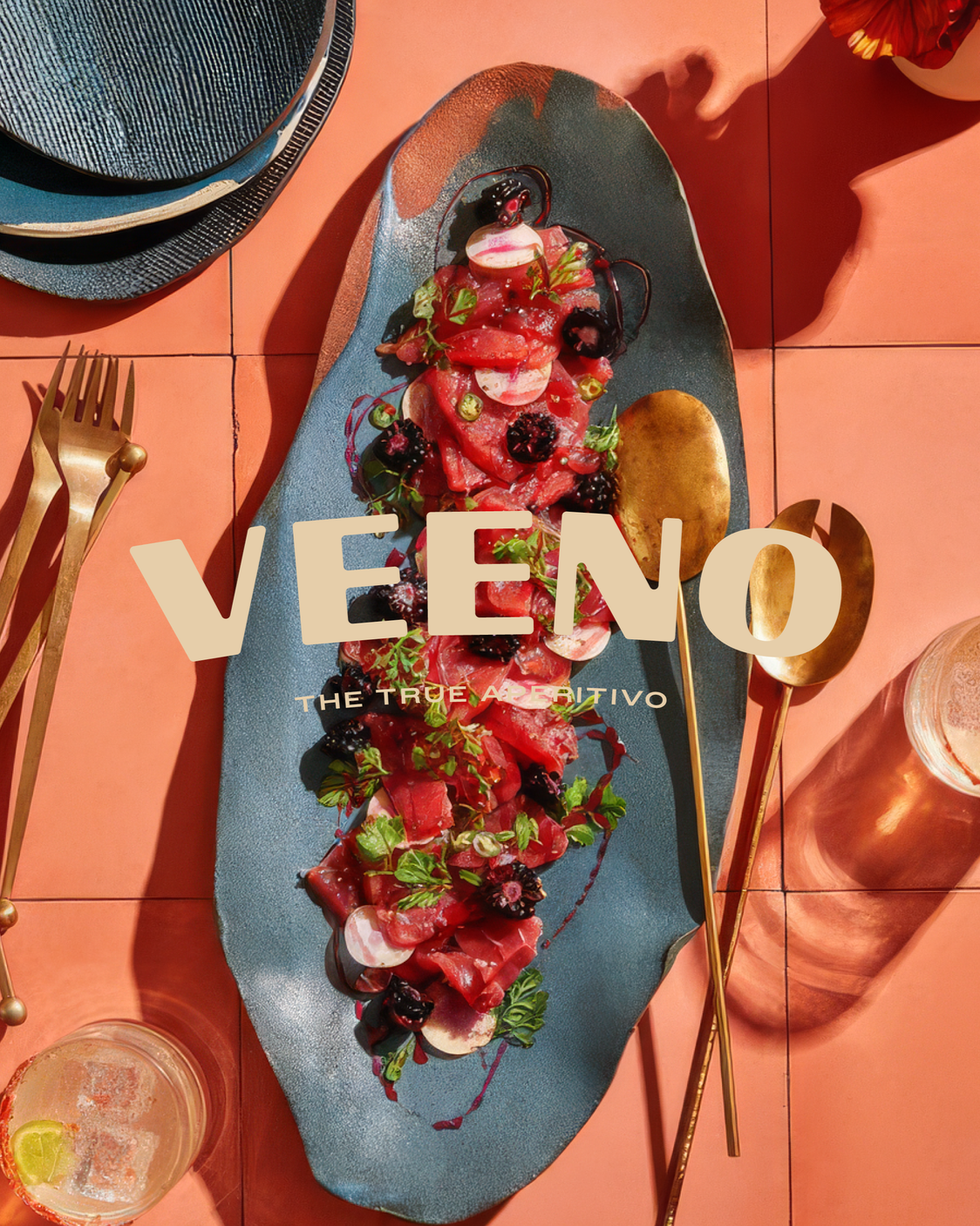 A plated dish of sliced cured meat, fresh herbs, berries, and thin sliced vegetables on a blue oval plate, surrounded by gold utensils, glasses, and additional plates, on a pink tiled table. Overlay text reads 'V EE NO THE TRUE AD ERITIVO'.