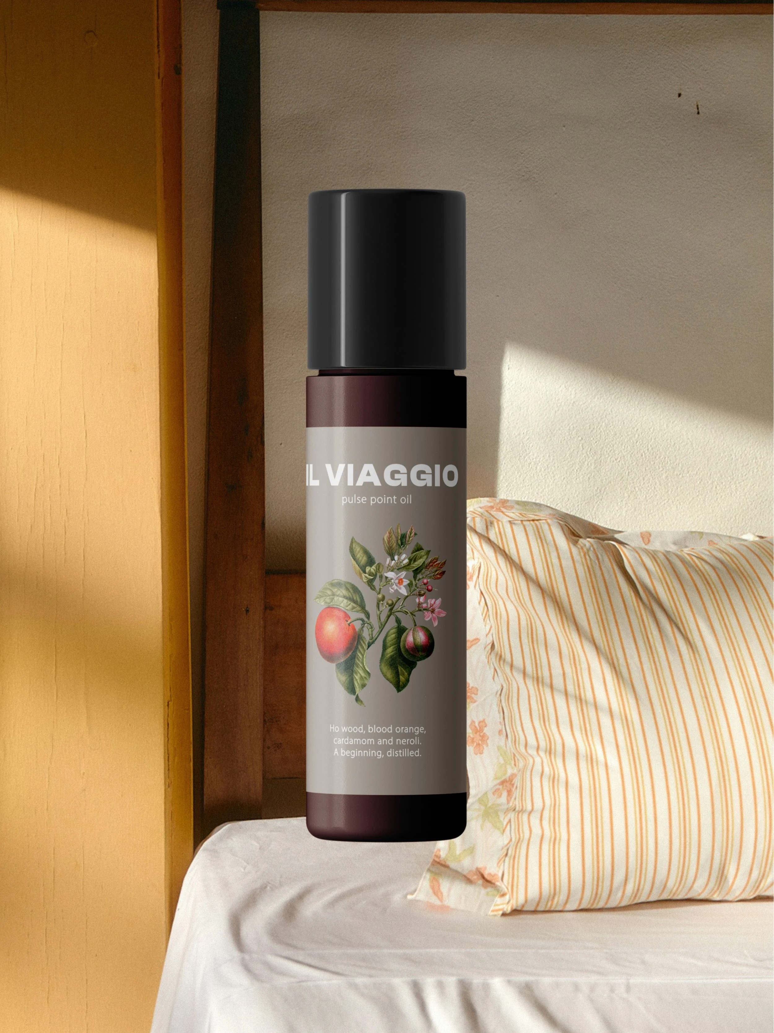 A cylindrical bottle of IL VIAGGIO pulse point oil with illustrations of fruits and flowers, placed on a bed with a striped pillow in a room.