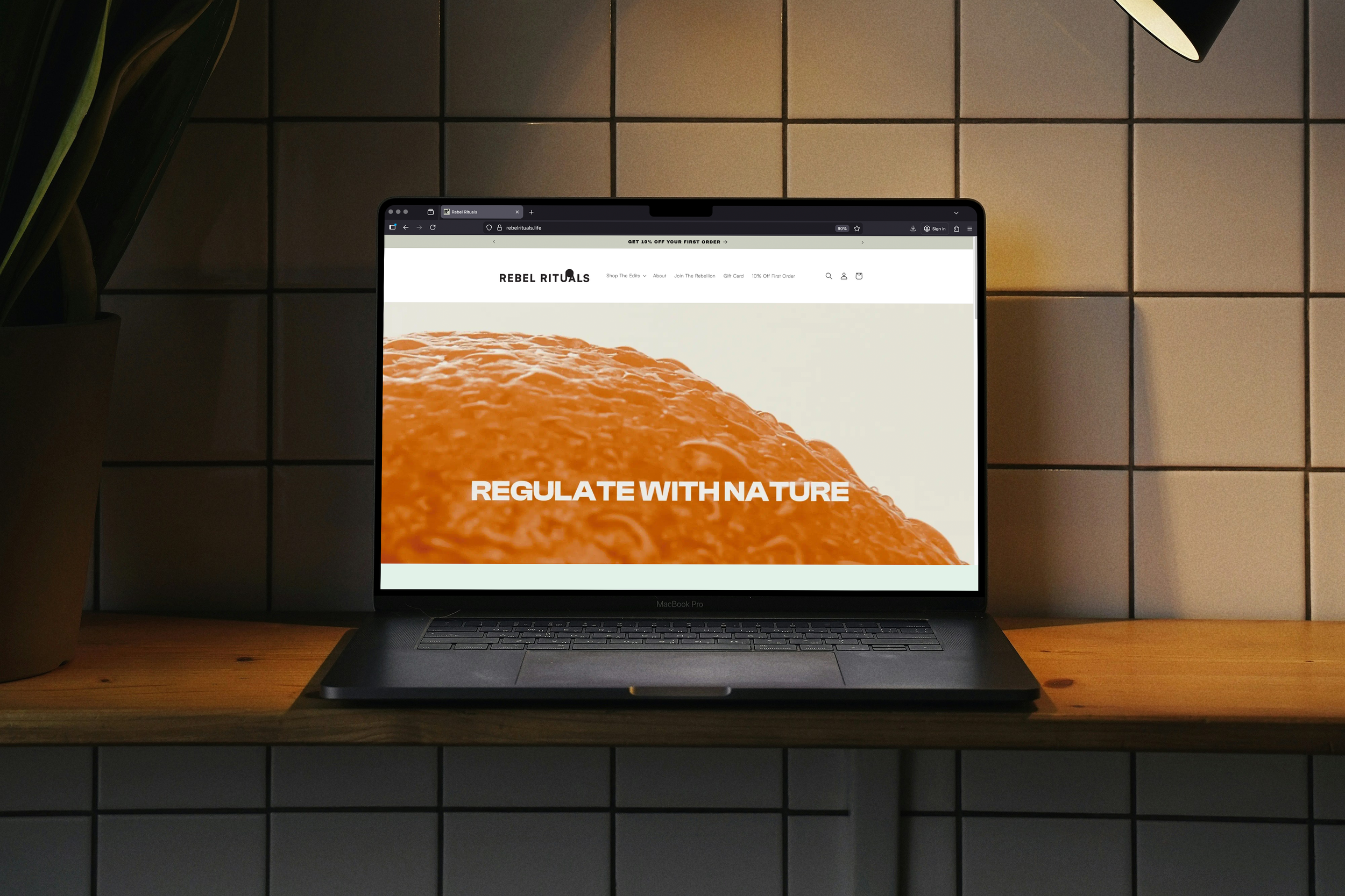 A MacBook Pro laptop on a wooden desk displaying the Rebel Rituals website with an orange textured background and the text "Regulate With Nature".